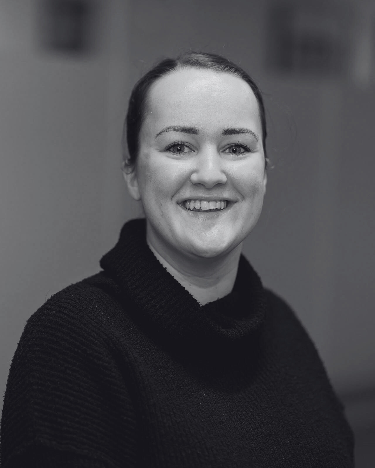 Black and white portrait of a smiling woman wearing a dark sweater, looking at the camera with a plain background.