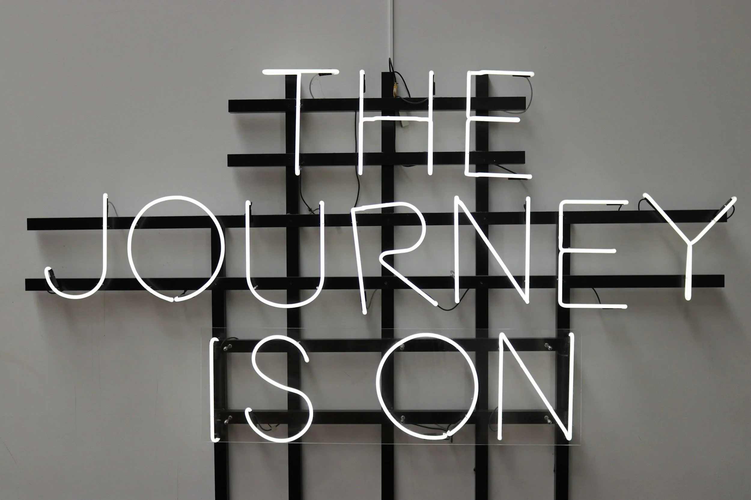 A neon sign artistically displaying the words 'THE JOURNEY' in white neon lights mounted on black horizontal and vertical bars against a plain light-colored wall.