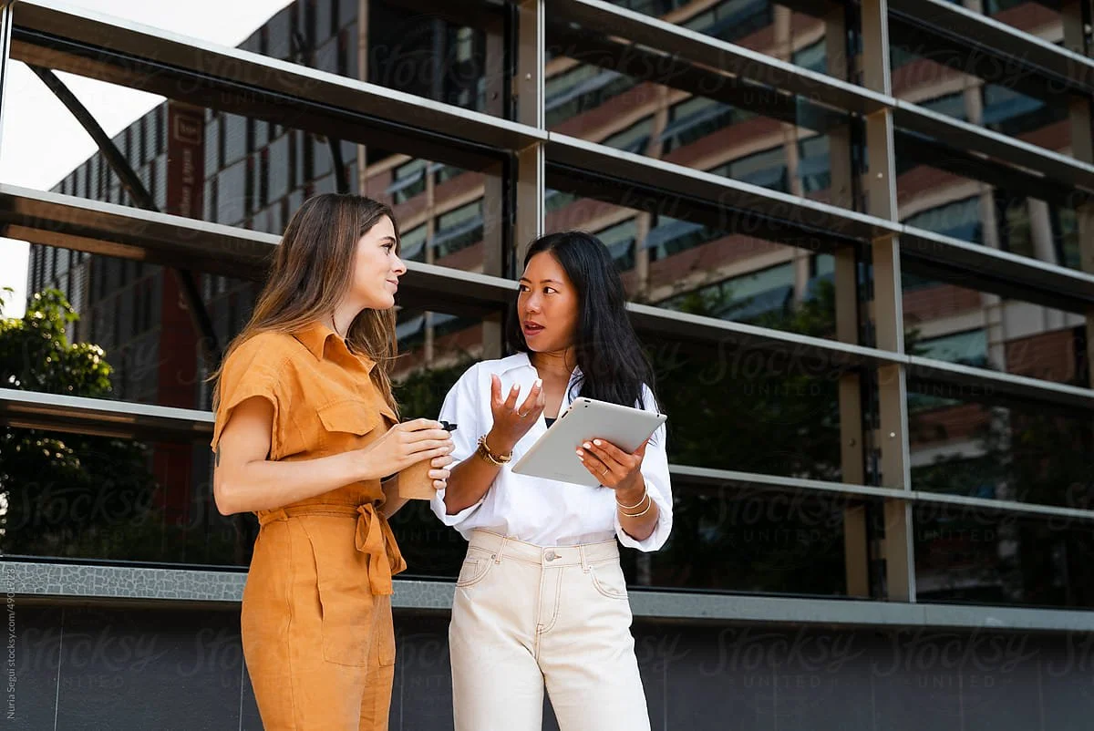 Two women are standing outside in front of a modern glass building, engaged in conversation. One woman is holding a tablet, and the other is holding a pen and a notebook.