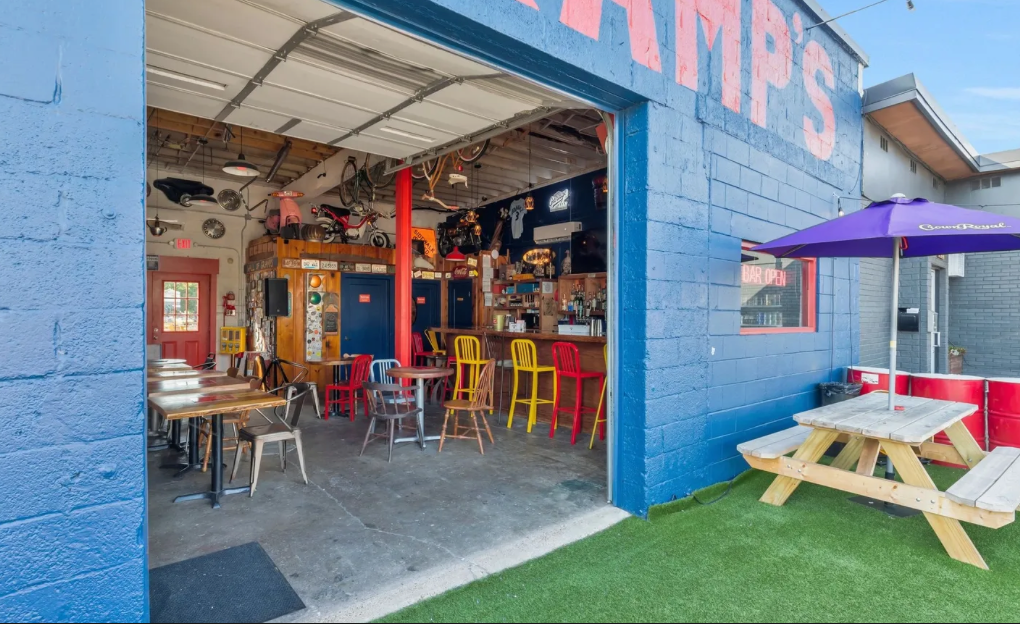 View of a colorful bar and patio area, featuring wooden tables, a purple umbrella, and vibrant chairs inside and outside.