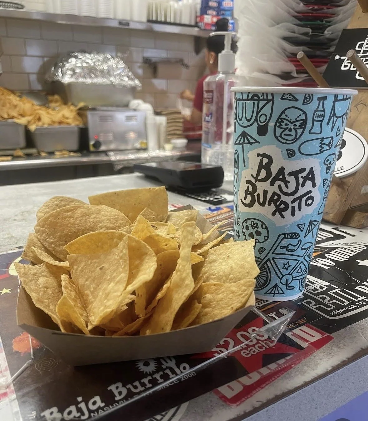 A paper tray of potato chips and a large cup with a Baja Burrito logo on it, on a counter at a Mexican restaurant.