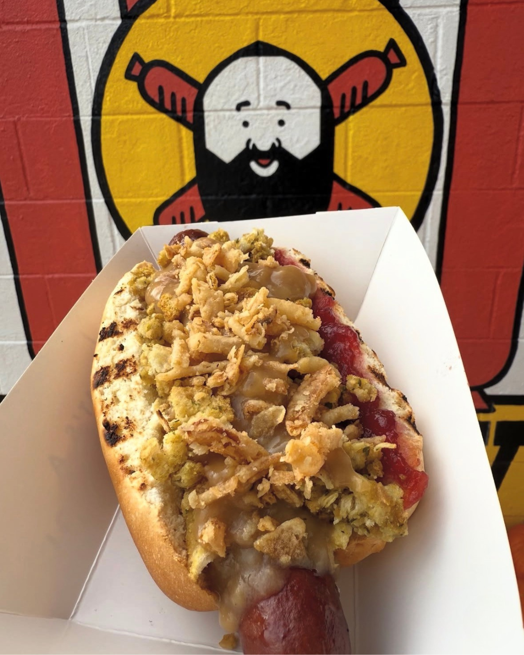 Hot dog with fried onions, relish, and ketchup in a paper holder in front of a restaurant wall featuring a cartoon chef logo.