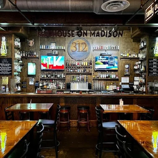 Interior of a bar named House on Madison with wooden tables and chairs, a bar counter with various bottles, two TVs displaying different channels, and TV screens on the walls.
