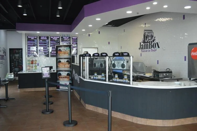 Inside JamBox Sandwich Shop, showing the counter, menu boards, and snack display, with a Coke machine on the right.