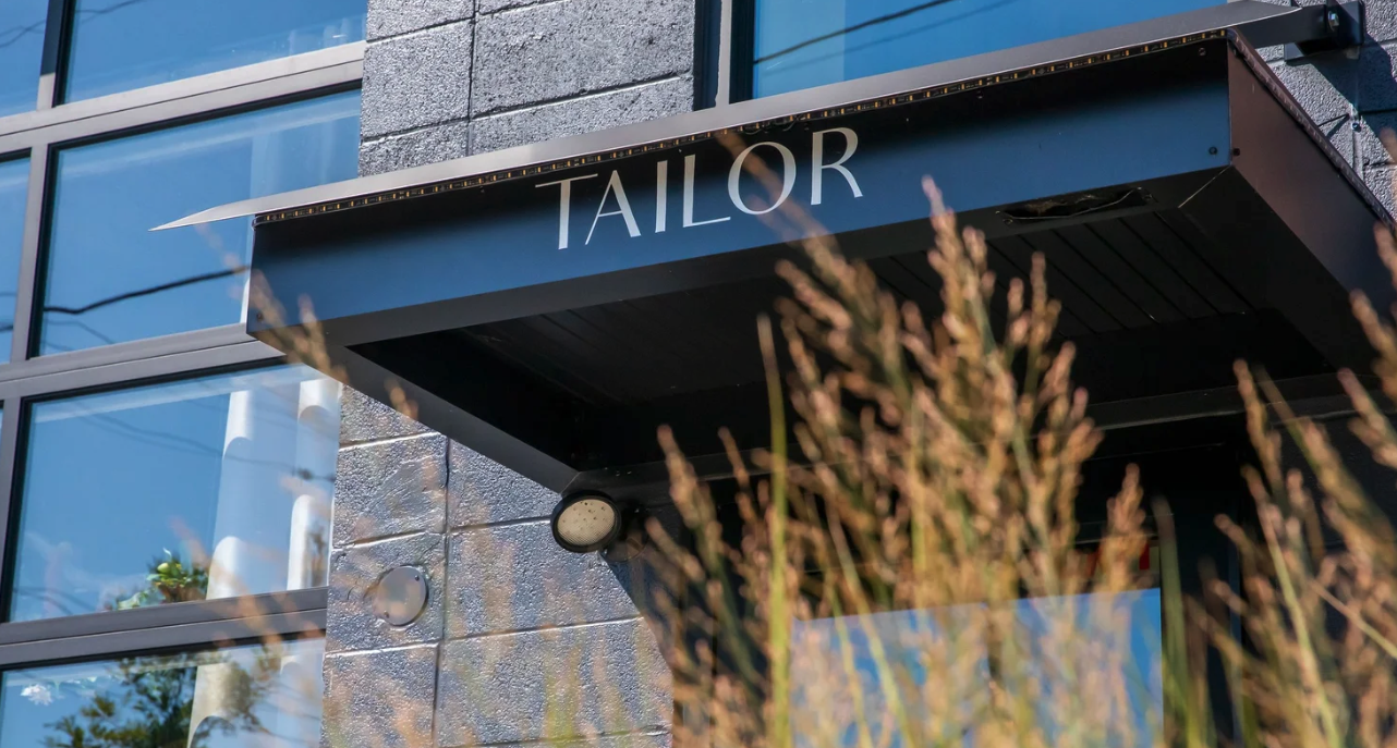 Exterior of a building with a black awning displaying the word 'TAYLOR' in white letters, glass windows reflecting the sky, and some plants in the foreground.