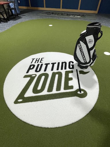 Indoor golf putting area with a green artificial turf, a large circular sign reading 'The Putting Zone,' and a golf bag with clubs.