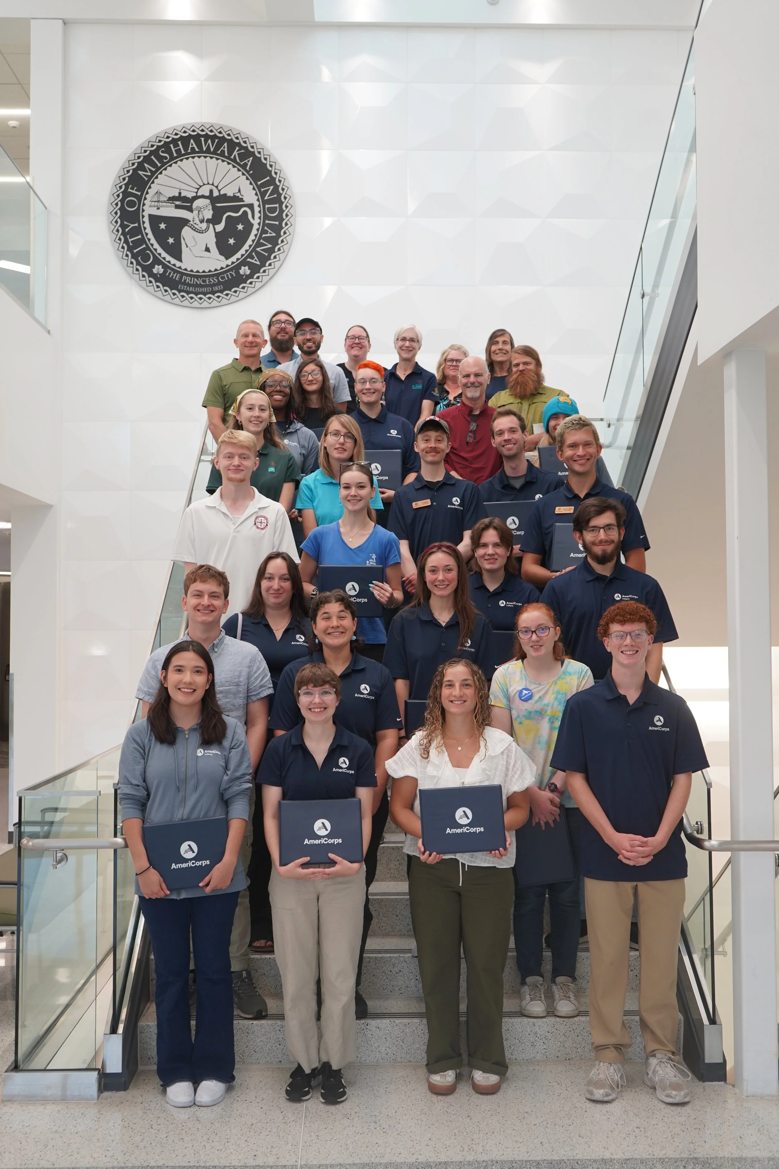 A large group of AmeriCorps members stand on a staircase for a group photo with their certificates.