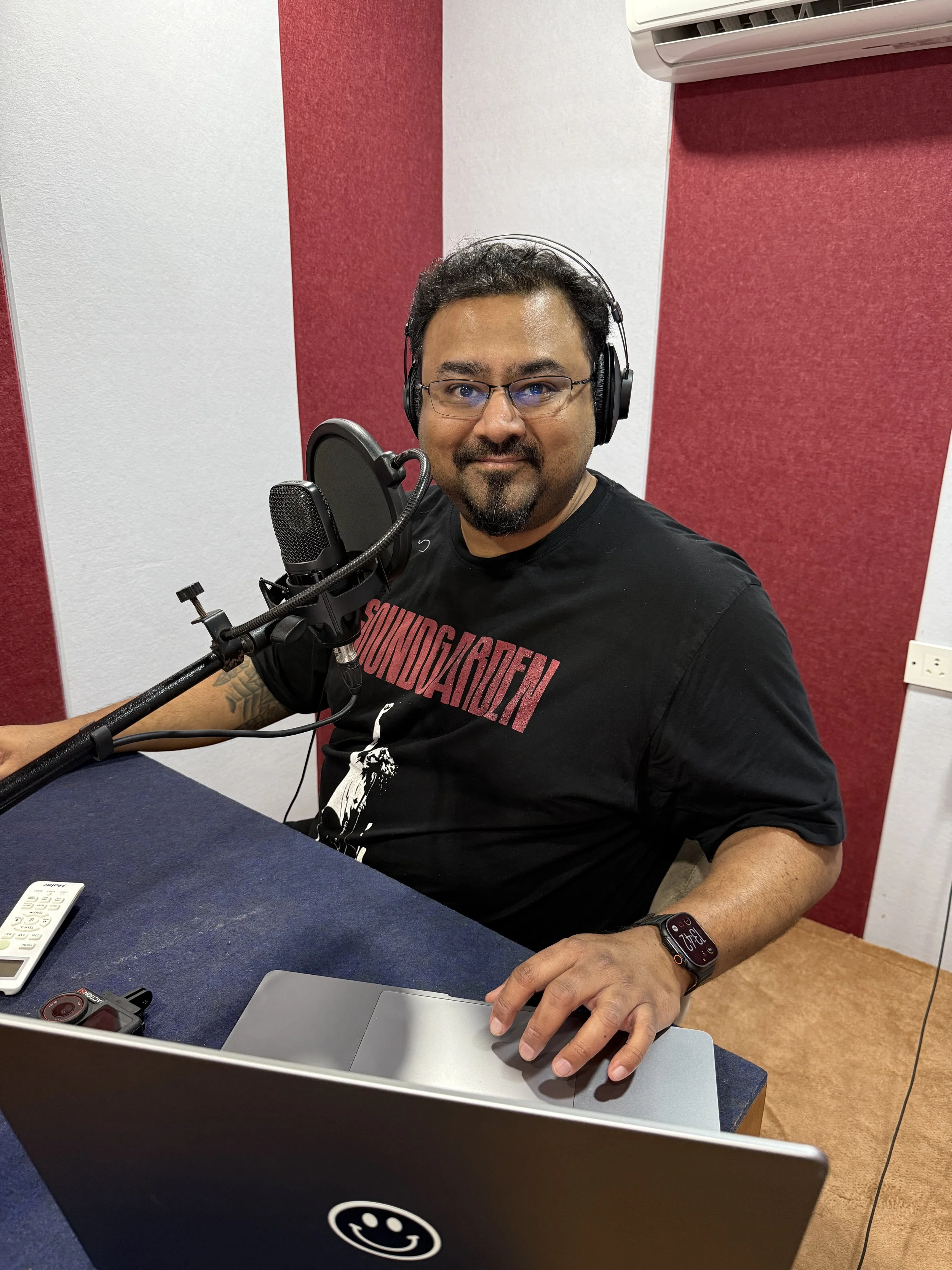 A man with glasses and a beard, sitting at a table in a recording studio or radio station, wearing headphones, with a microphone in front of him, touching a laptop or notepad, and a remote control and a camera on the table, with acoustic foam panels on the wall behind him.