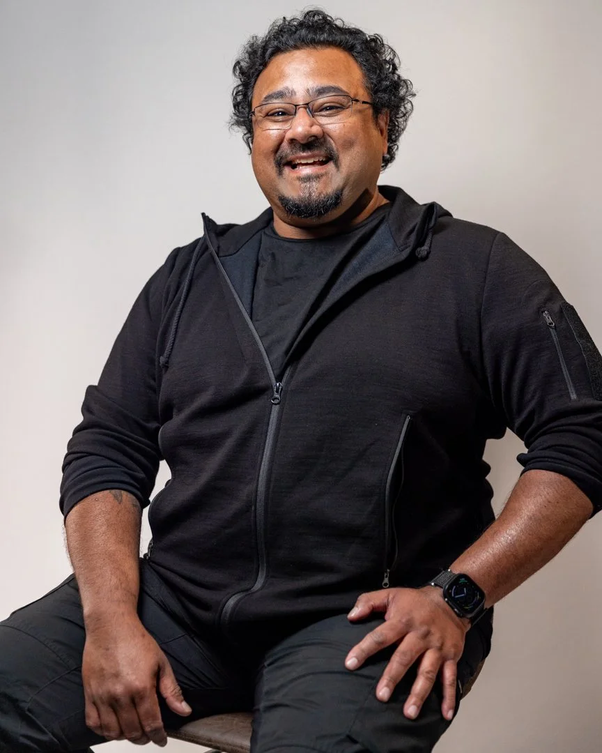 A man with curly black hair, glasses, and a beard, smiling while sitting on a chair, wearing a black hoodie, black t-shirt, black pants, and a black smartwatch.