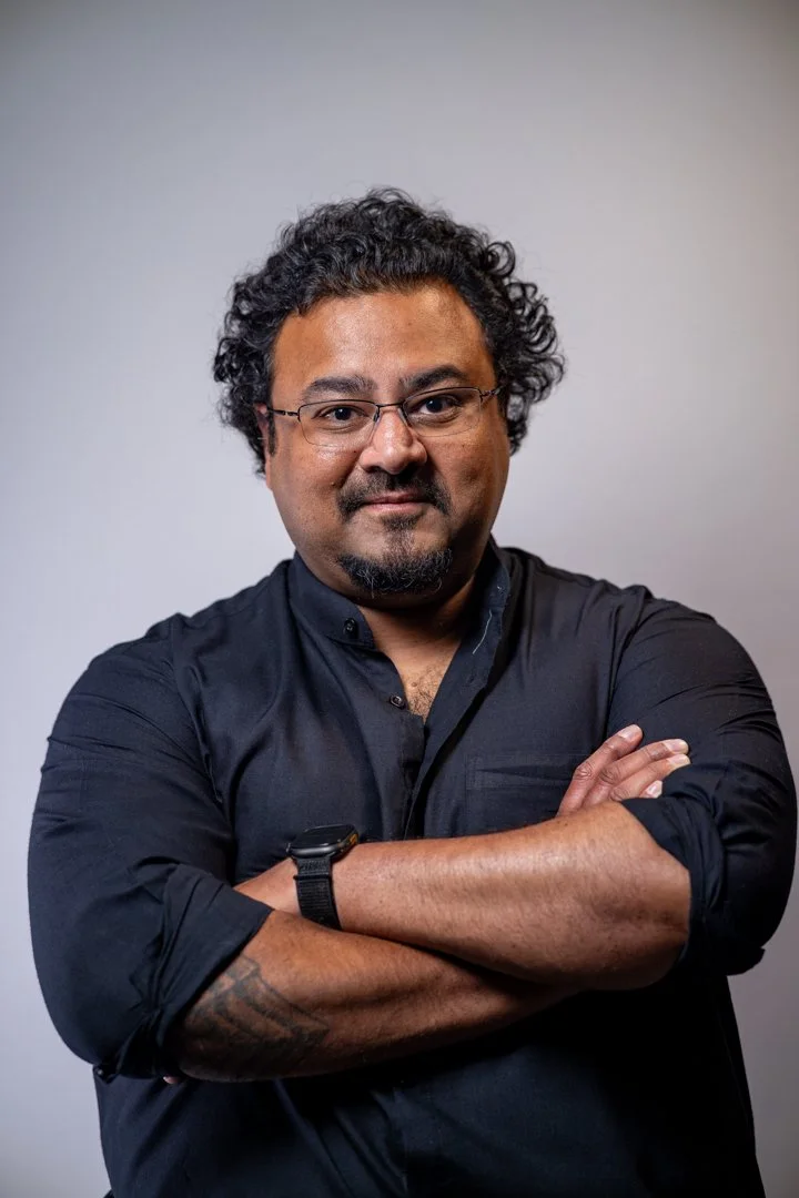 A man with curly hair and glasses, wearing a black shirt, standing with arms crossed against a neutral background.