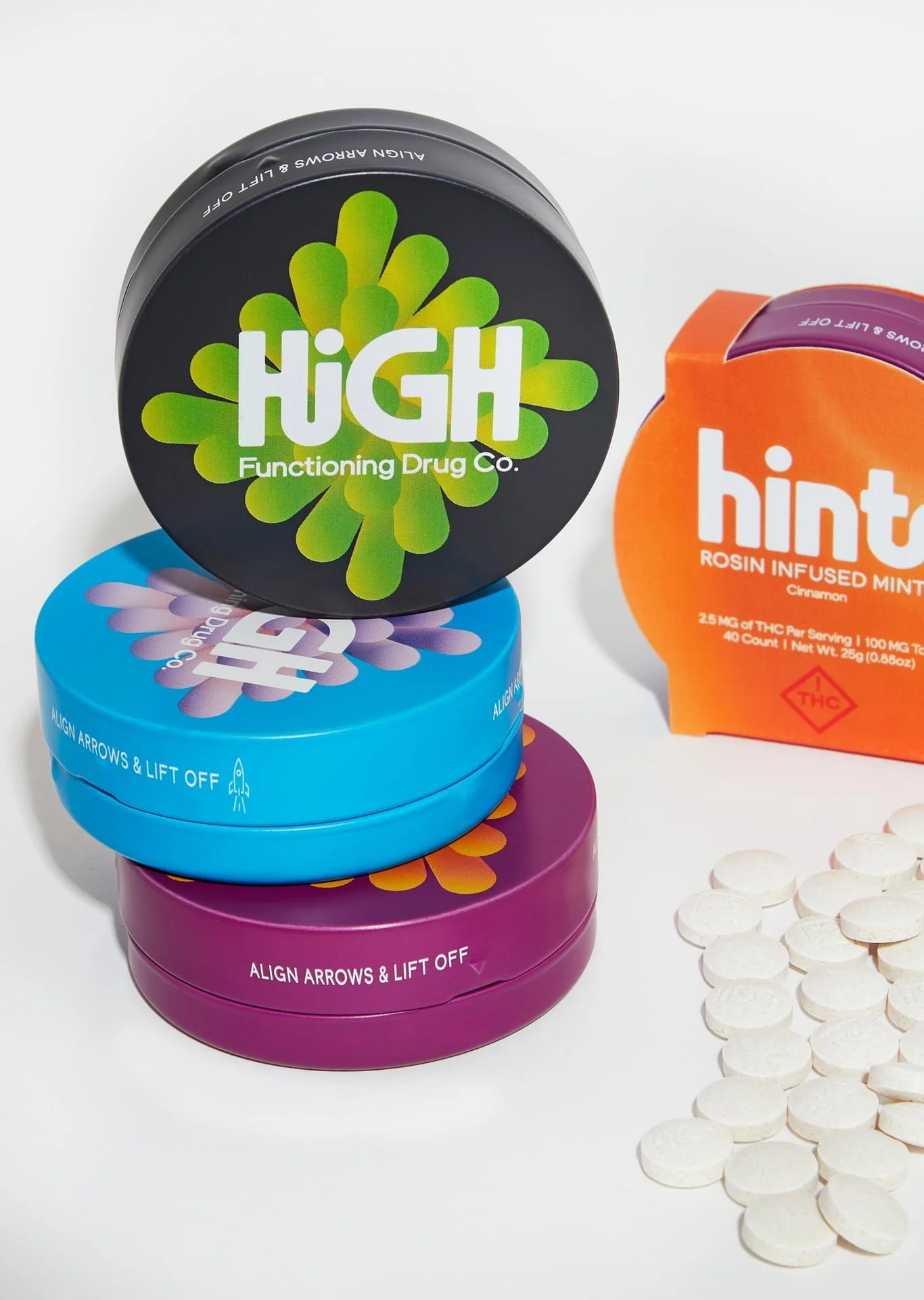 Stacked colorful cannabis-themed tins with a blue tin on bottom, purple in the middle, and black on top, with a box of orange and purple mints to the right and scattered white tablets on the right.