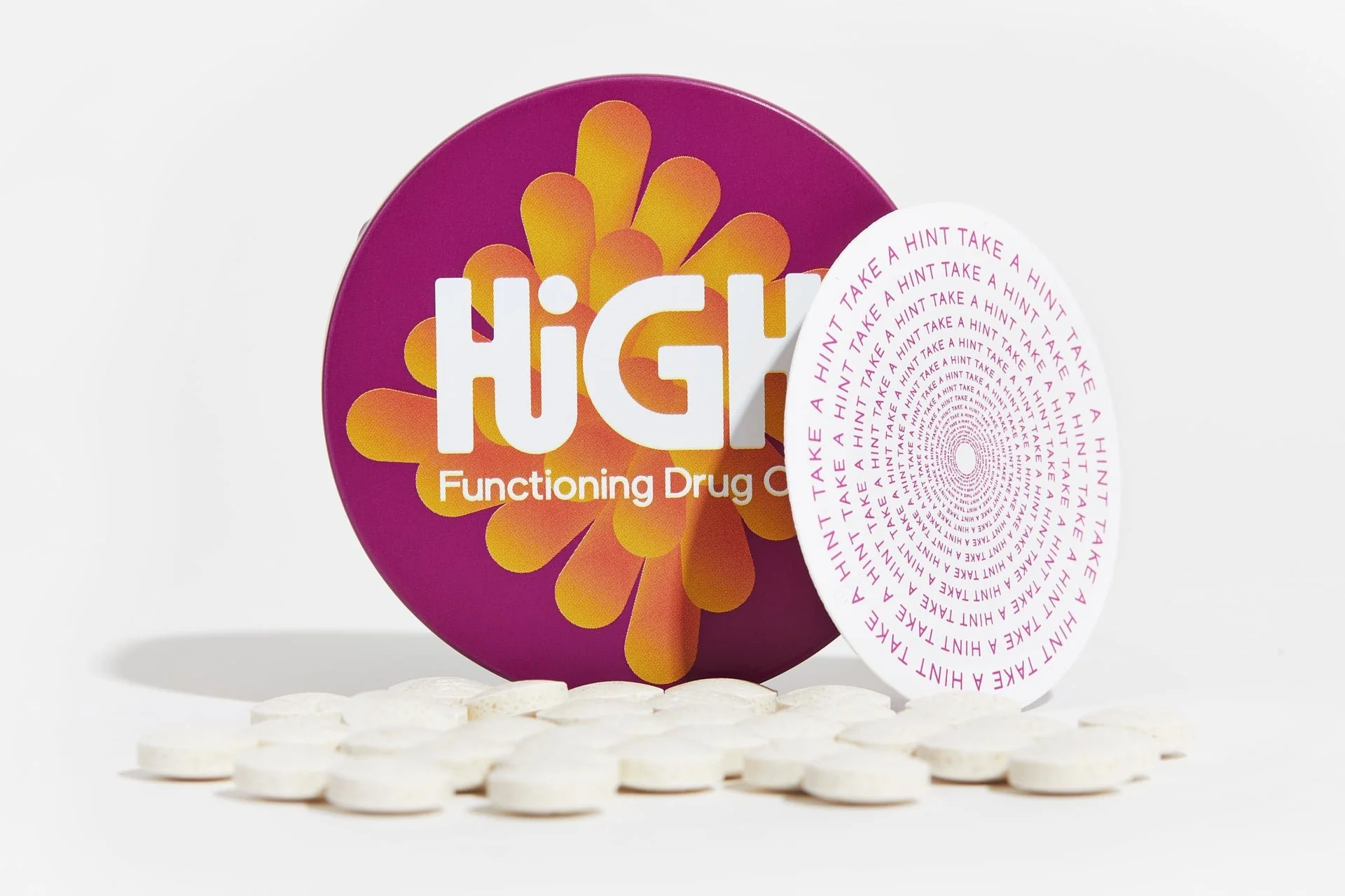 A purple round tin labeled 'High Functioning Drug' with orange floral design, surrounded by white pills, and a white card with pink text spiraling inward that says 'Take a Hint' repeatedly.