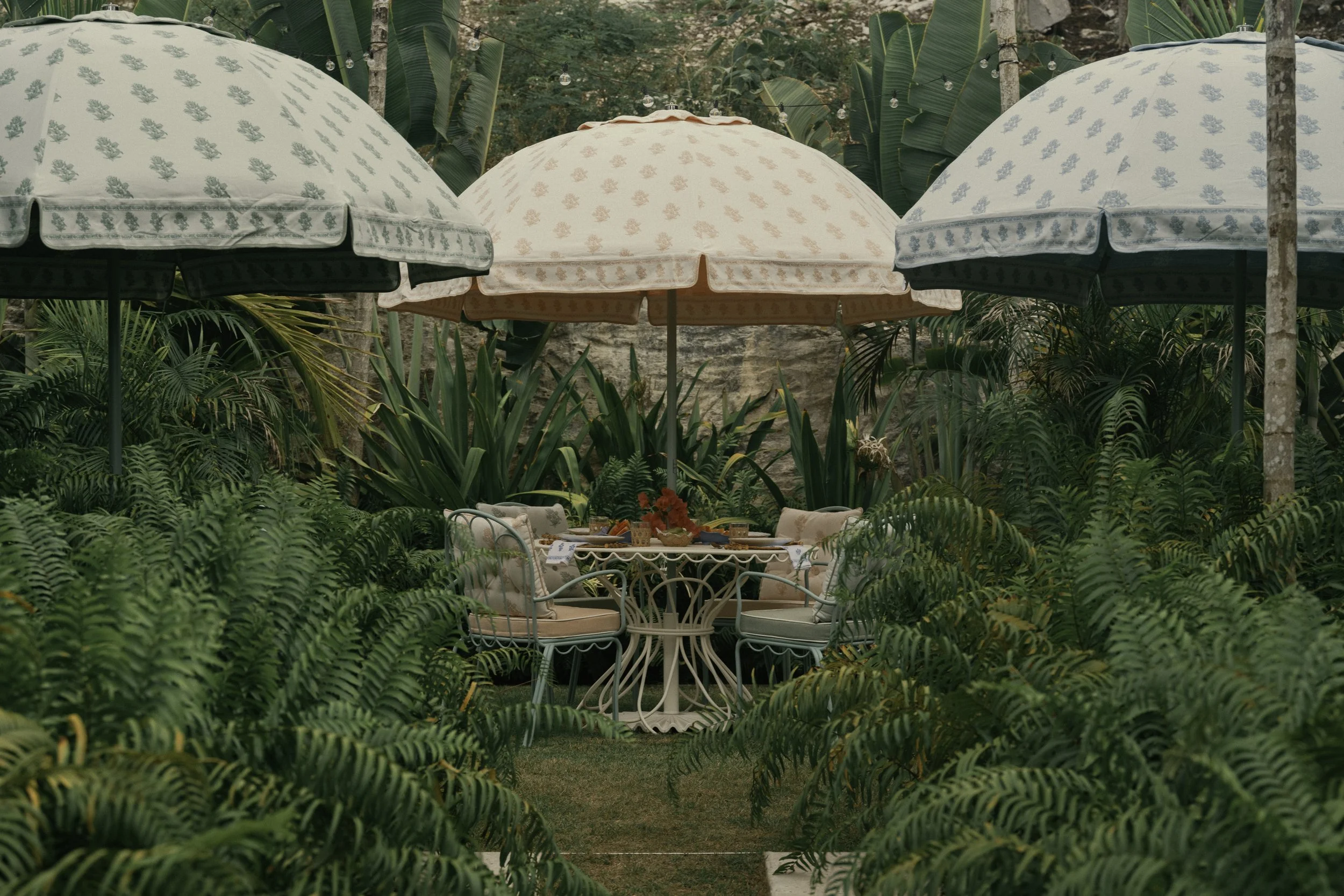 Outdoor dining area with a round table and cushioned chairs, surrounded by lush greenery including palm leaves and ferns, with three large umbrellas providing shade.