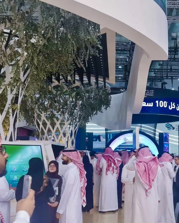 A group of men wearing traditional Saudi attire, including white thobes and red-and-white checkered shemaghs, gathered in an indoor exhibition or conference center with modern architectural features.