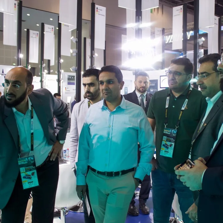 Group of men in business attire at a technology conference, engaging in discussion, with exhibition booths and digital displays in the background.