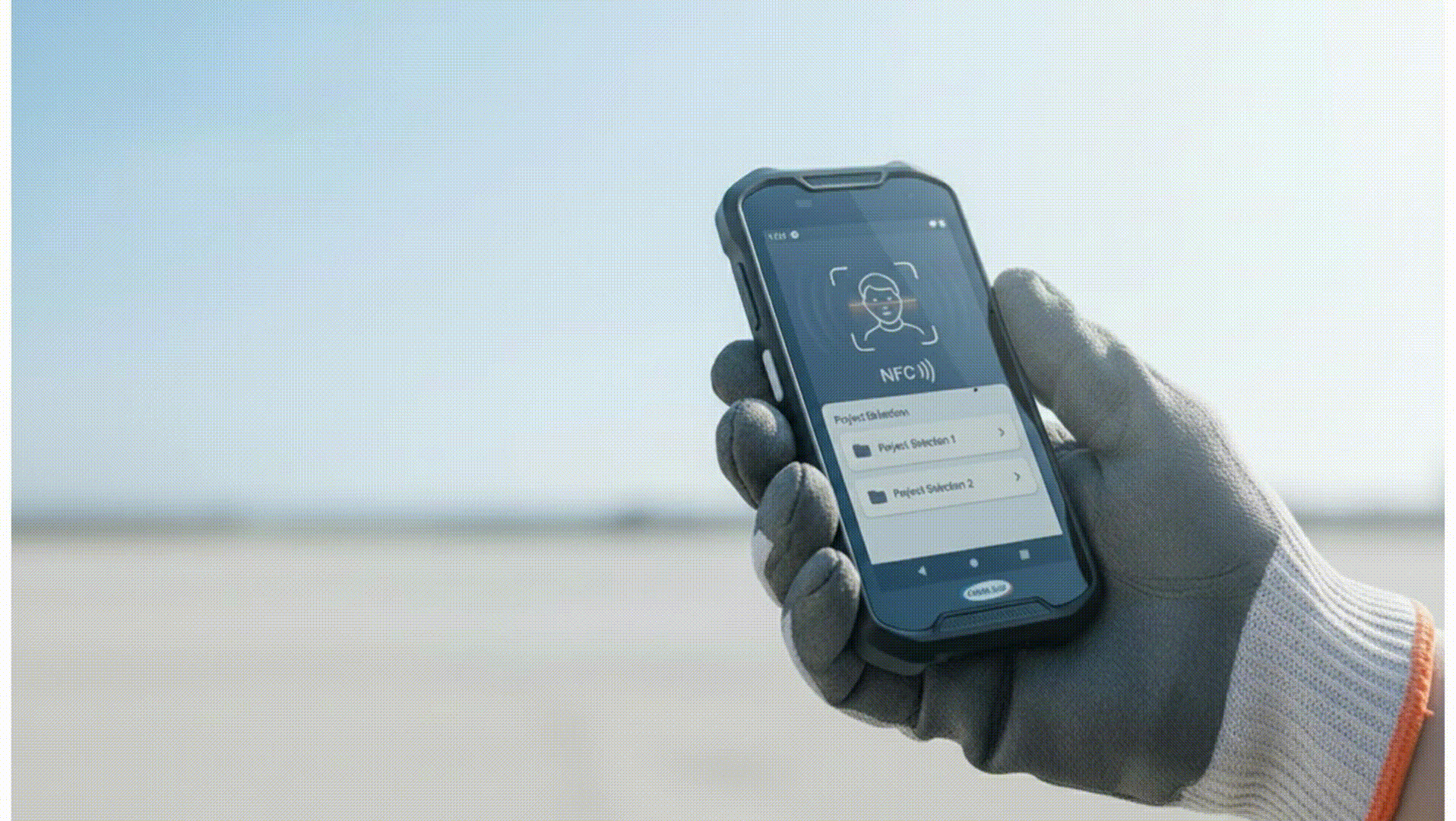 Eine Person mit Arbeitshandschuhen hält ein robustes Smartphone in der Hand, auf dem eine NFC-Zahlungs-App geöffnet ist; im Bild sind zwei Projektordner zu sehen; die Aufnahme wurde im Freien vor dem Hintergrund eines strahlend blauen Himmels gemacht.