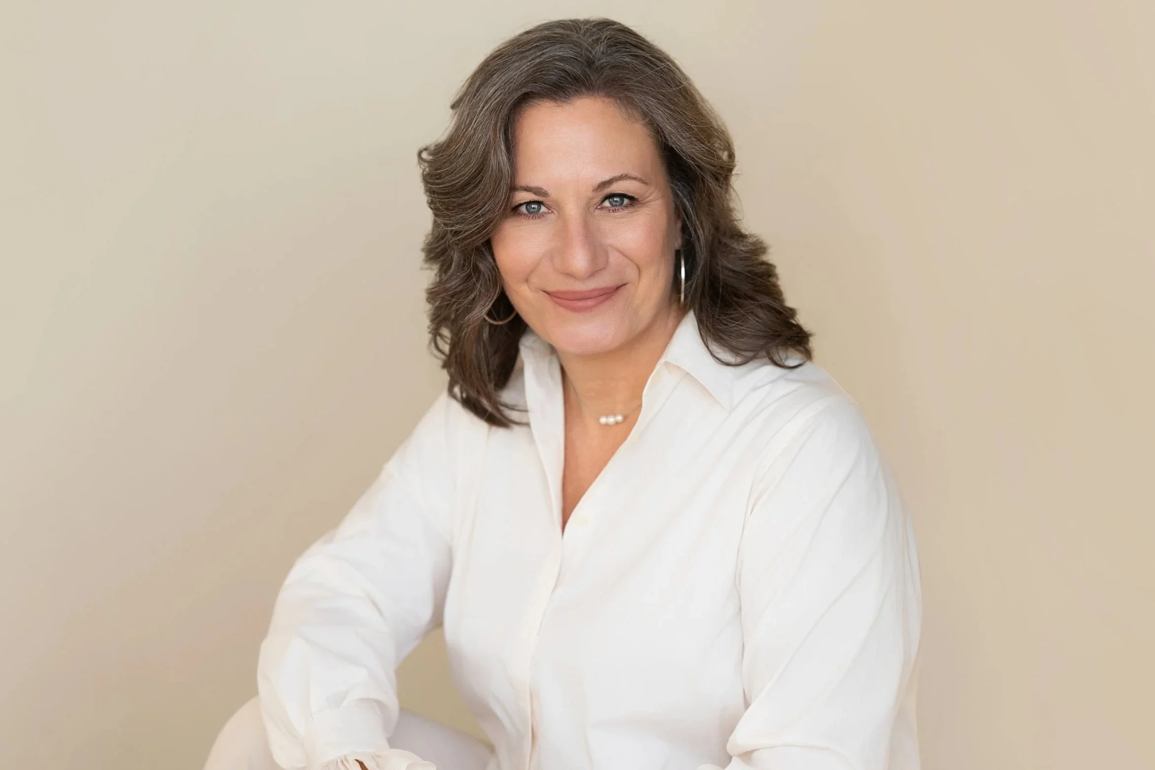 Femme aux cheveux bruns bouclés, portant une chemise blanche, souriant, contre un fond beige