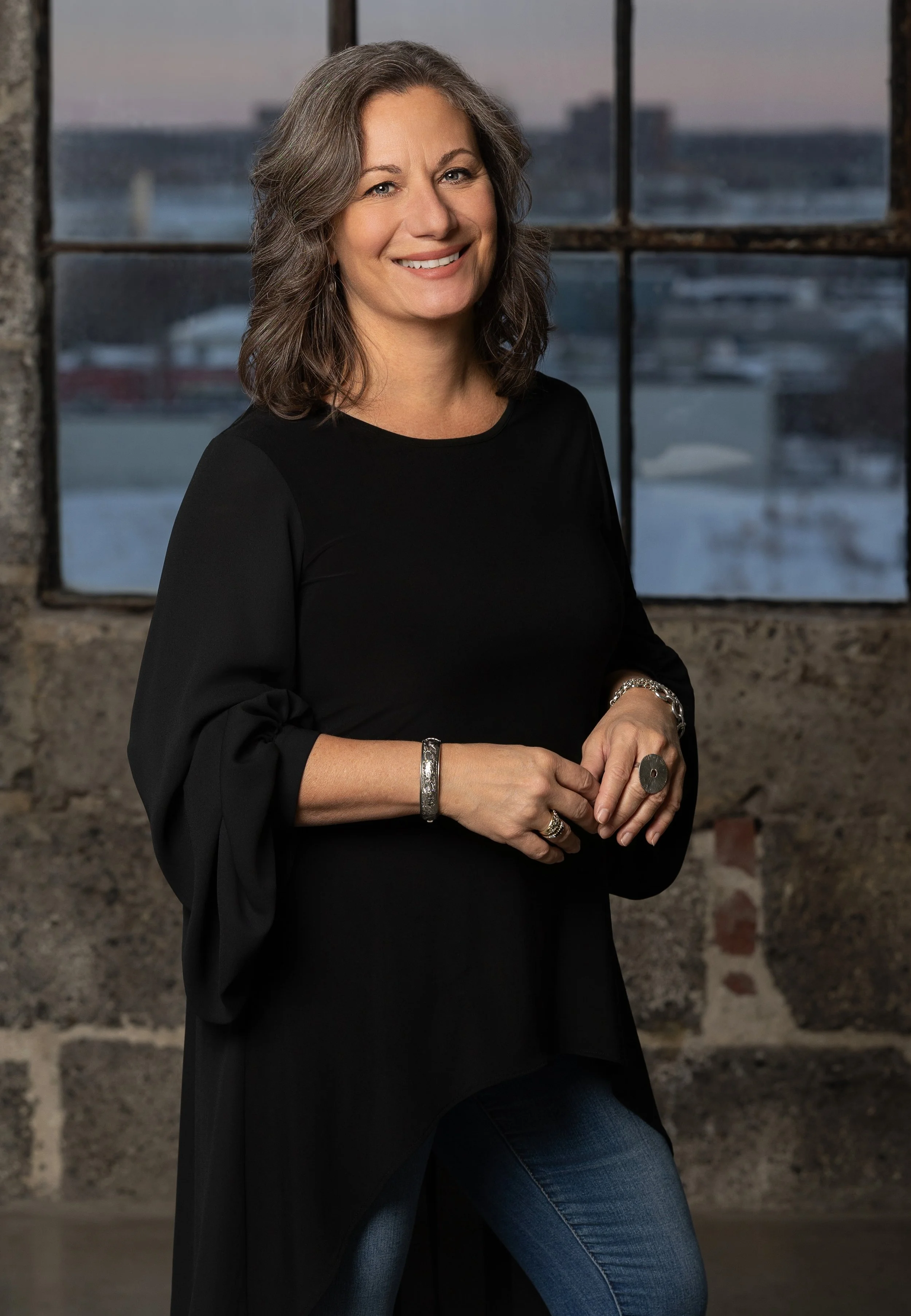 Femme souriante en tenue noire, debout devant une fenêtre avec vue sur le port.