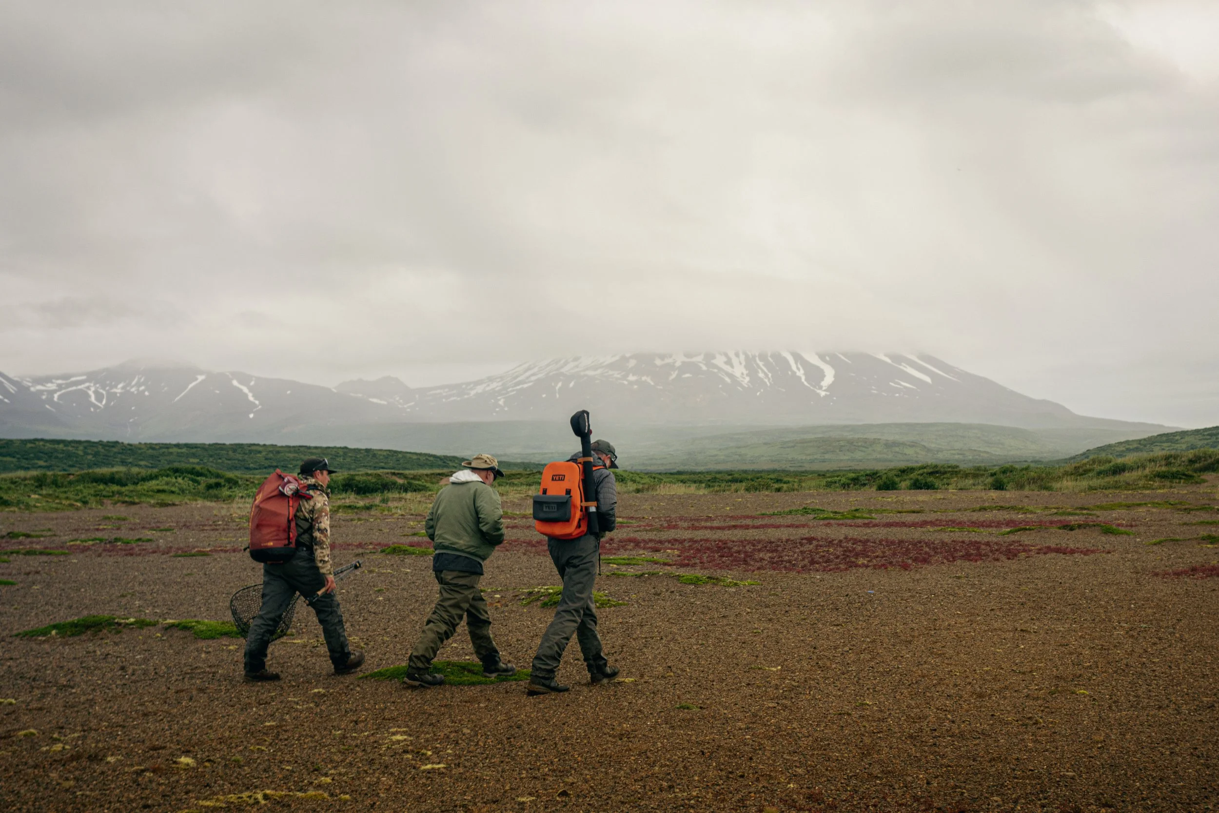 Three people walking across a barren landscape with a distant snow-capped mountain in the background.