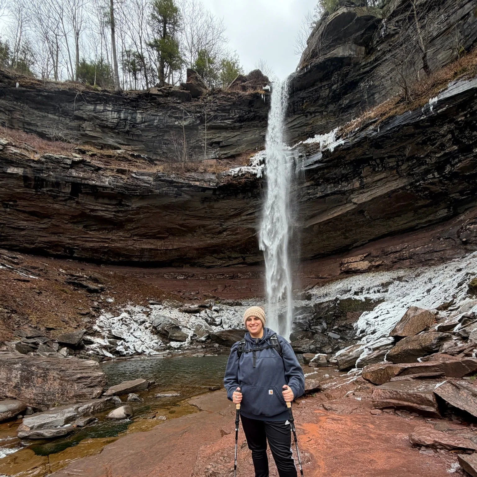 A woman in outdoor gear holding trekking poles stands in front of a waterfall surrounded by rocks and trees.
