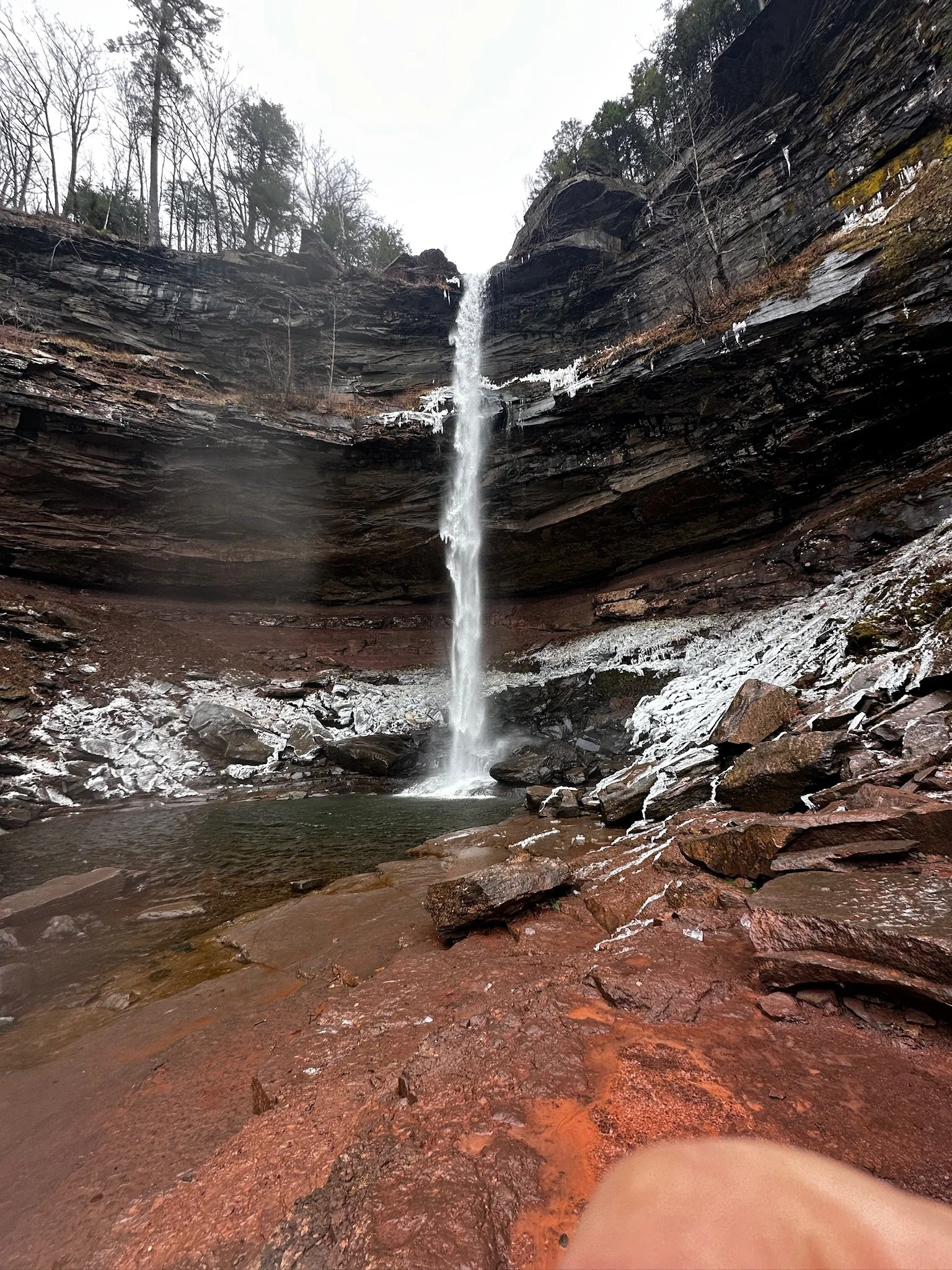 A narrow waterfall cascades down a dark, rocky cliffside into a small pool below, with leafless trees at the top and icy patches on the rocks.
