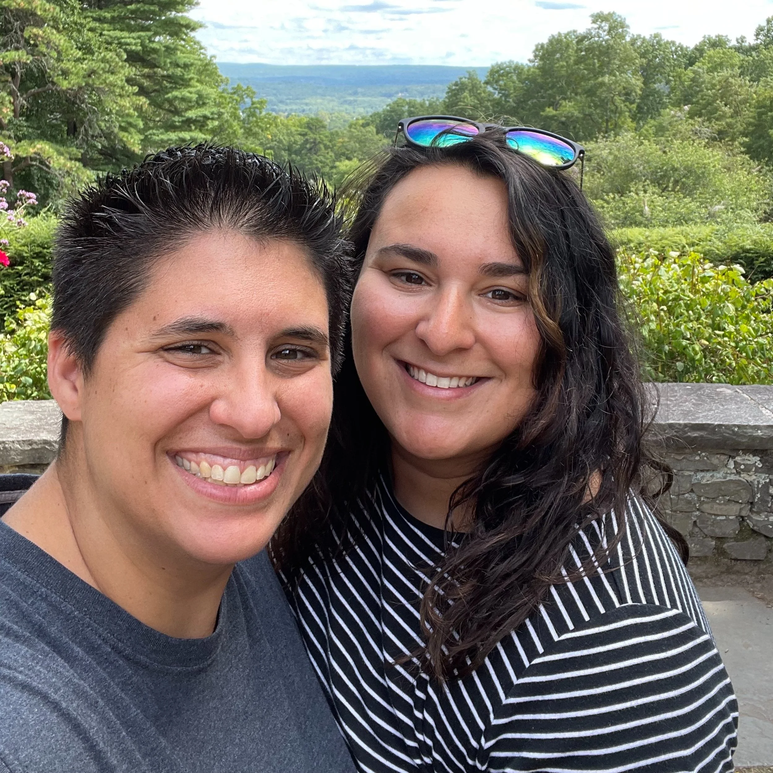 Two women smiling outdoors with a lush green landscape in the background, one with short dark hair and the other with long dark wavy hair and sunglasses on her head.