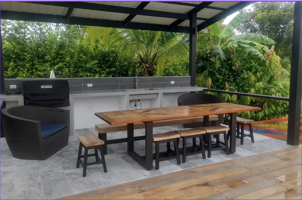 Outdoor covered patio with a wooden dining table, benches, and stools, a black wicker chair, a kitchen sink, and lush greenery including banana plants and flowering bushes.