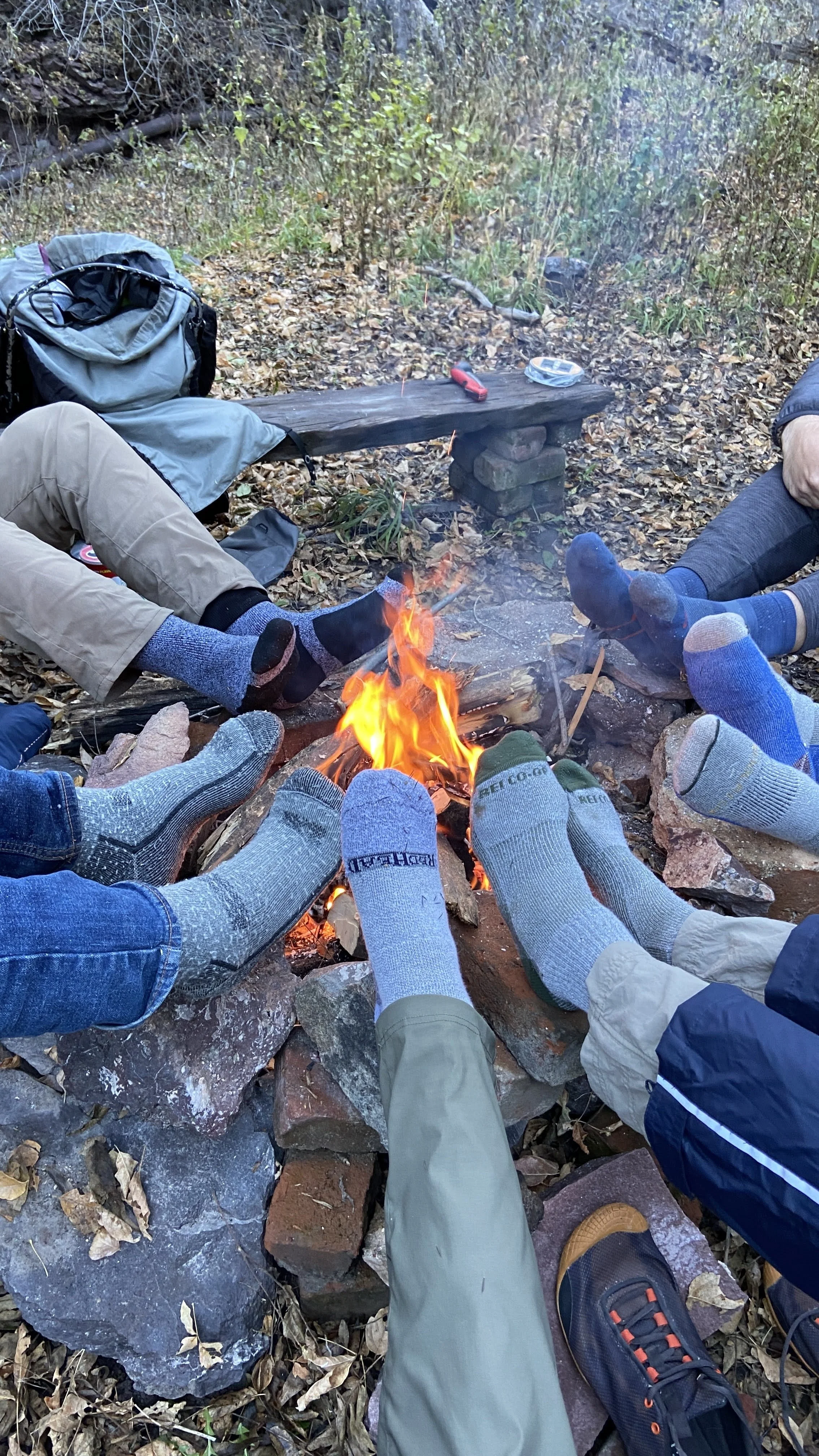 Group of people with socks on their feet sitting around a campfire in a wooded area with fallen leaves, a bench, and camping gear nearby.