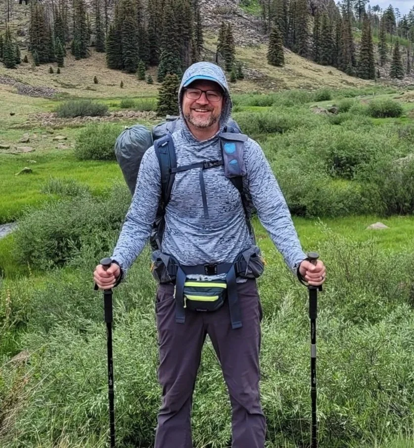 A man hiking in a lush green mountainous area, smiling, wearing a gray hooded jacket, glasses, a cap, backpack, and using trekking poles.