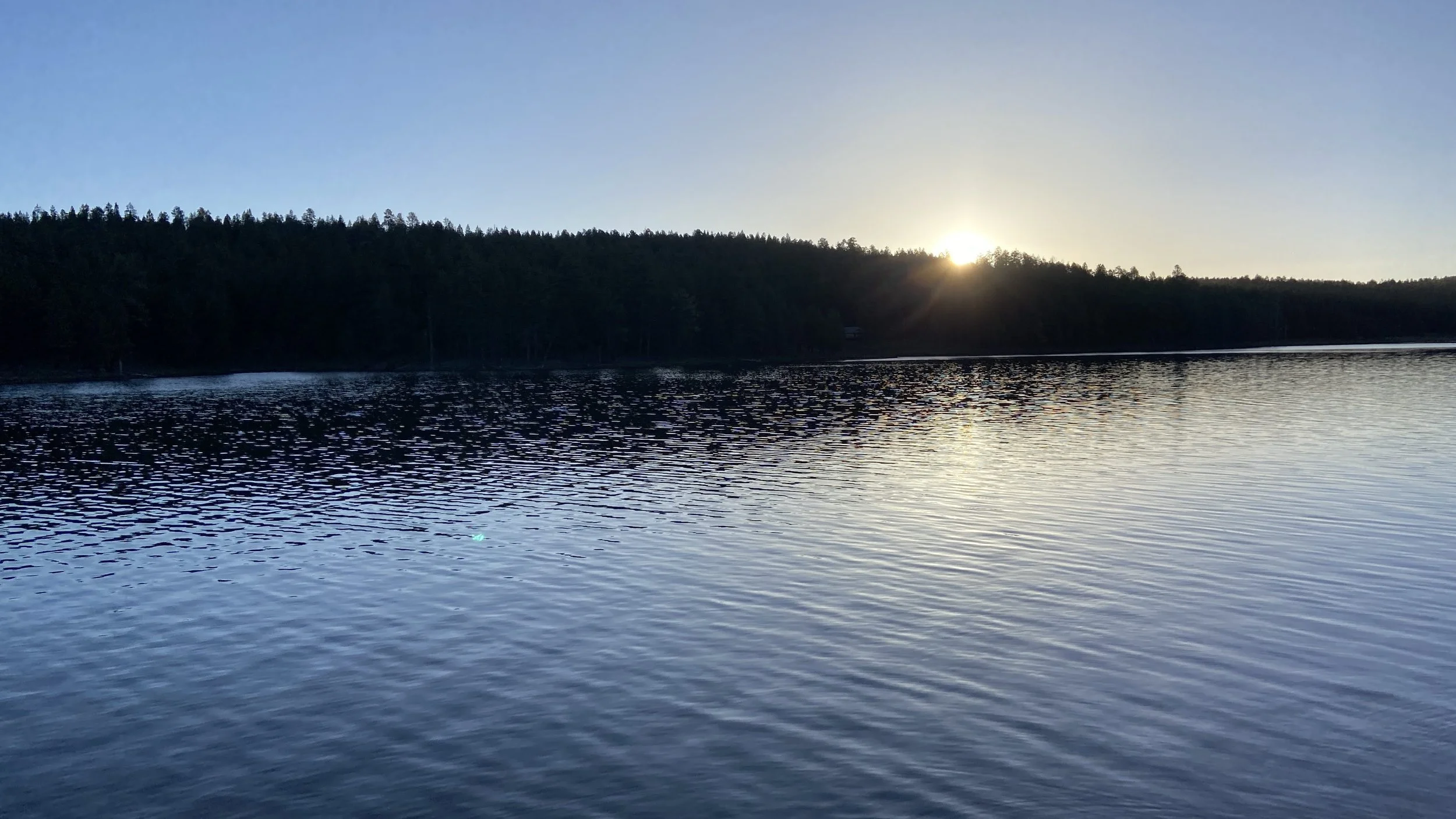 A tranquil lake at sunset with the sun partially behind a dark forested hill, reflecting light on the rippling water surface.