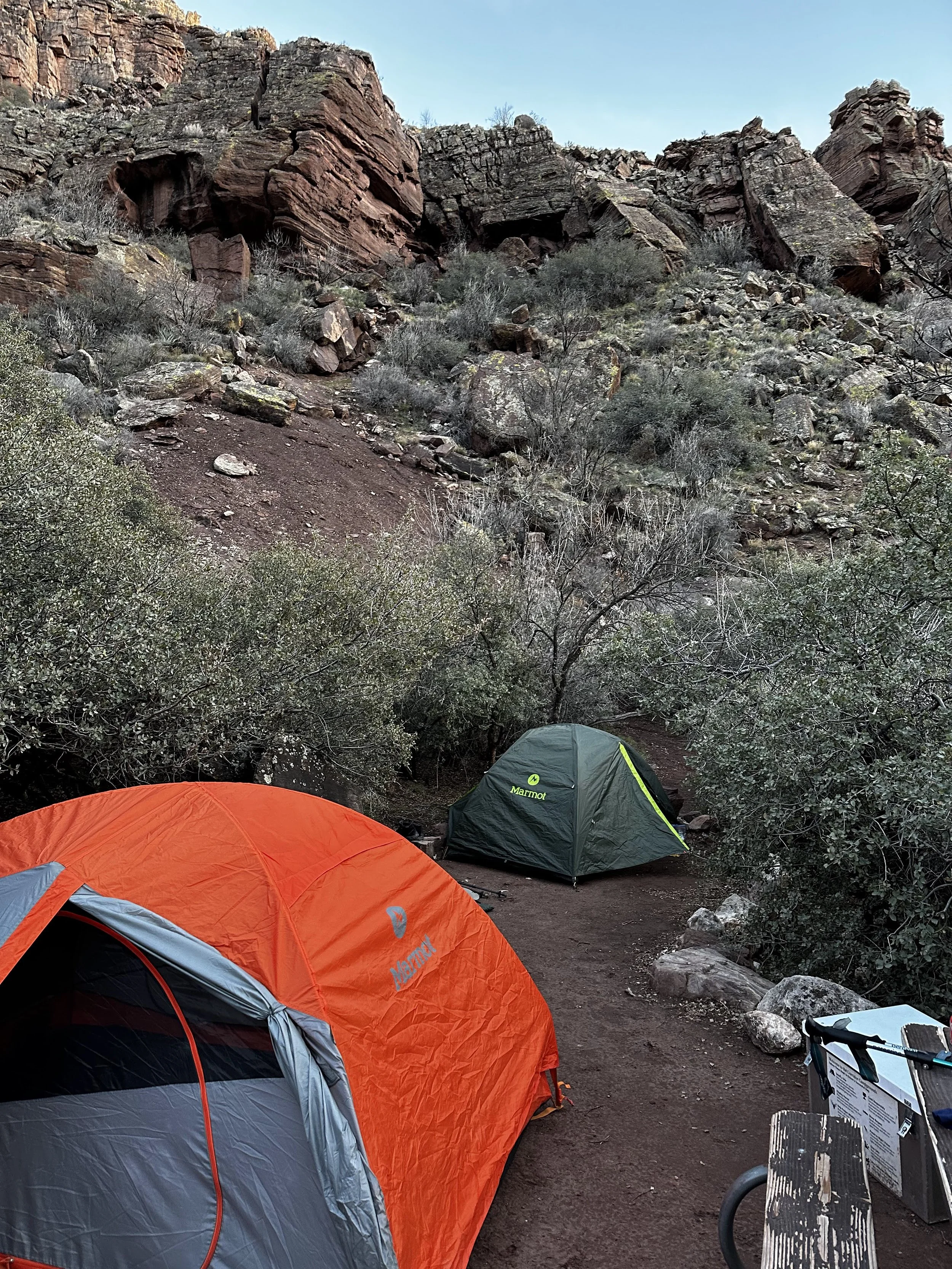 Camping tents set up on a dirt ground near rocky cliffs and sparse bushes, with a bench and some camping supplies visible in the foreground.