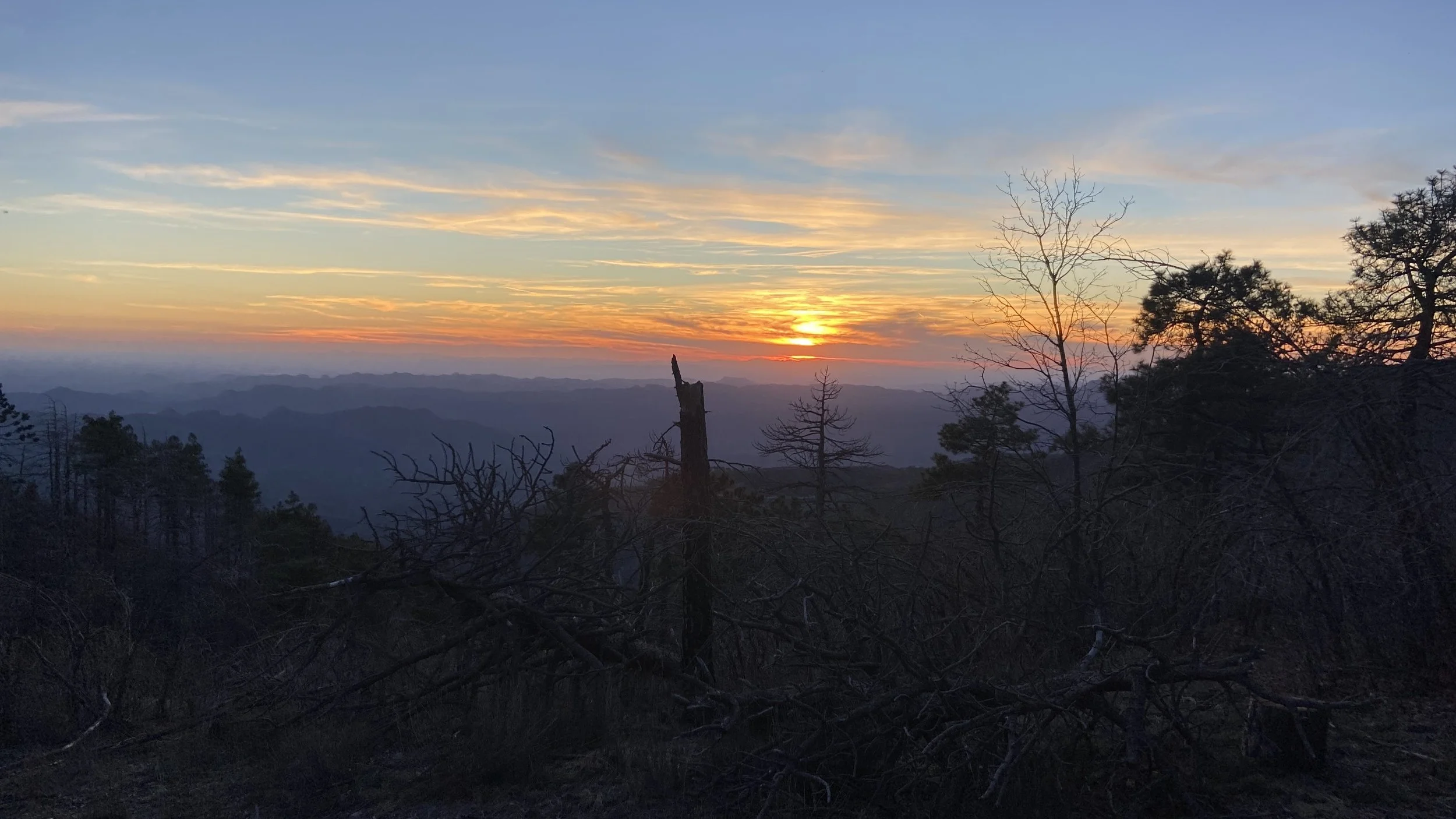 Sunset over a mountainous landscape with trees silhouetted against the colorful sky.
