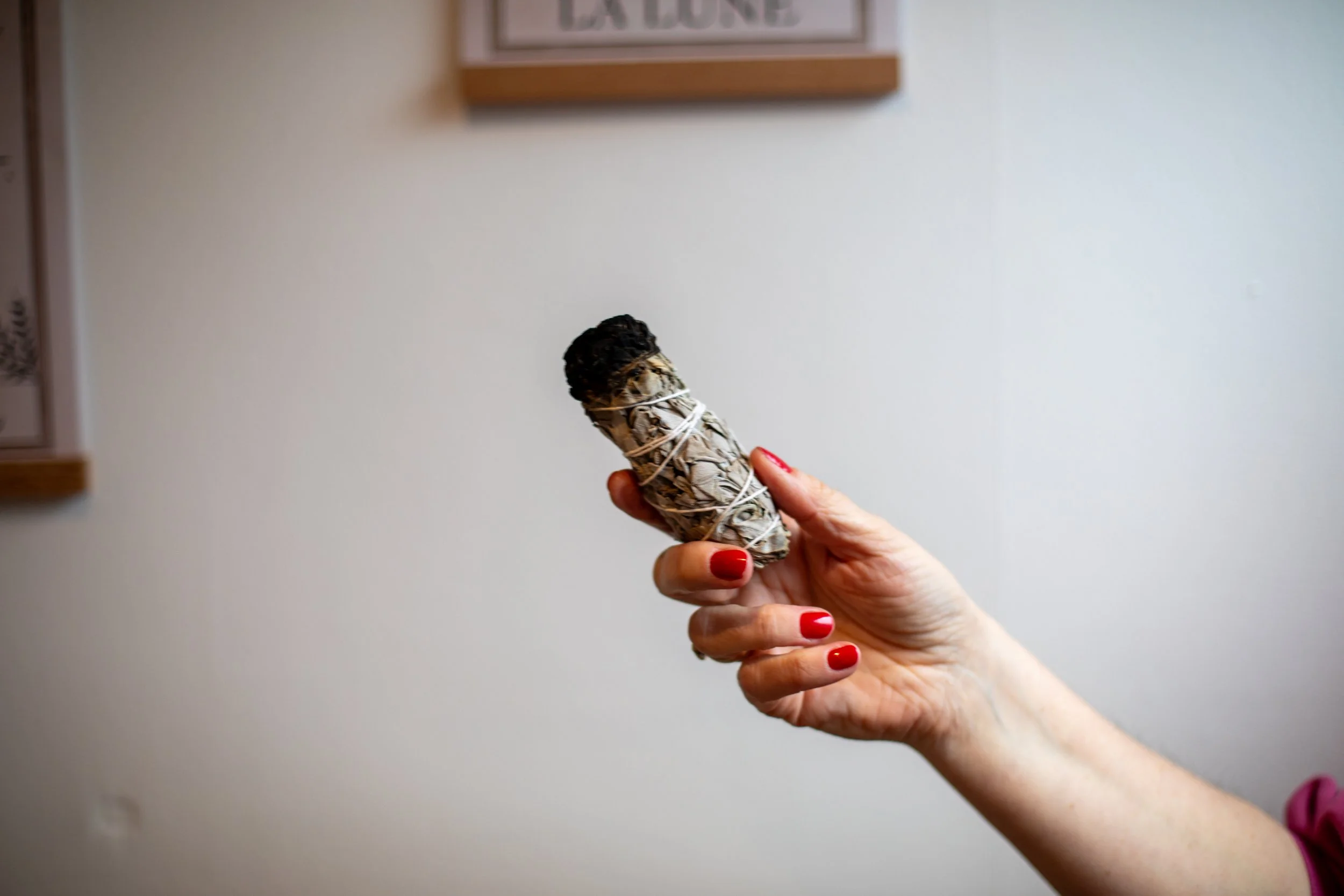A hand with red painted nails holding a smudged sage bundle tied with white string, with a black stone at the end, against a beige wall.