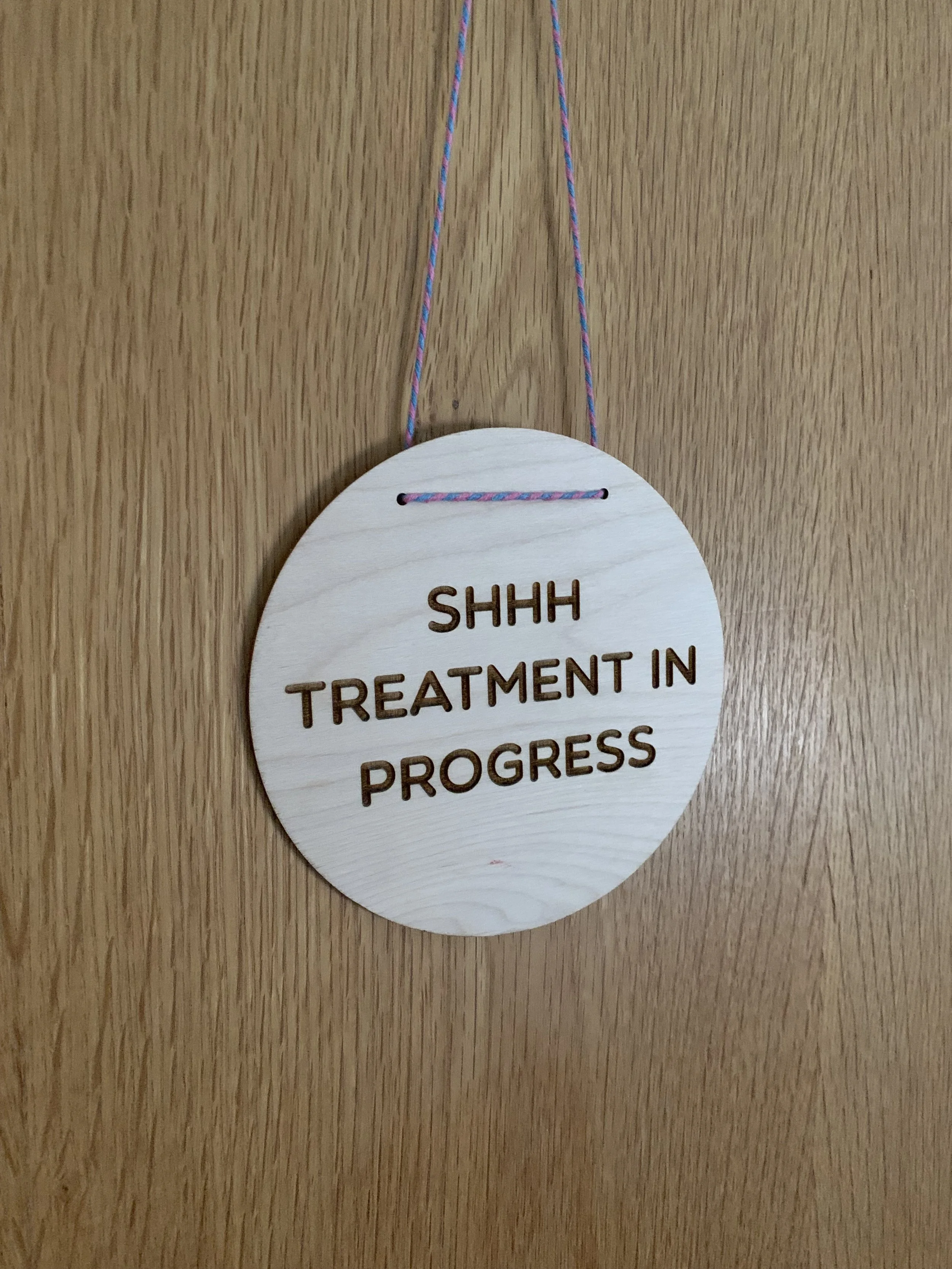 Wooden sign hanging on a door with a multicolored string, reading "SHHH TREATMENT IN PROGRESS."