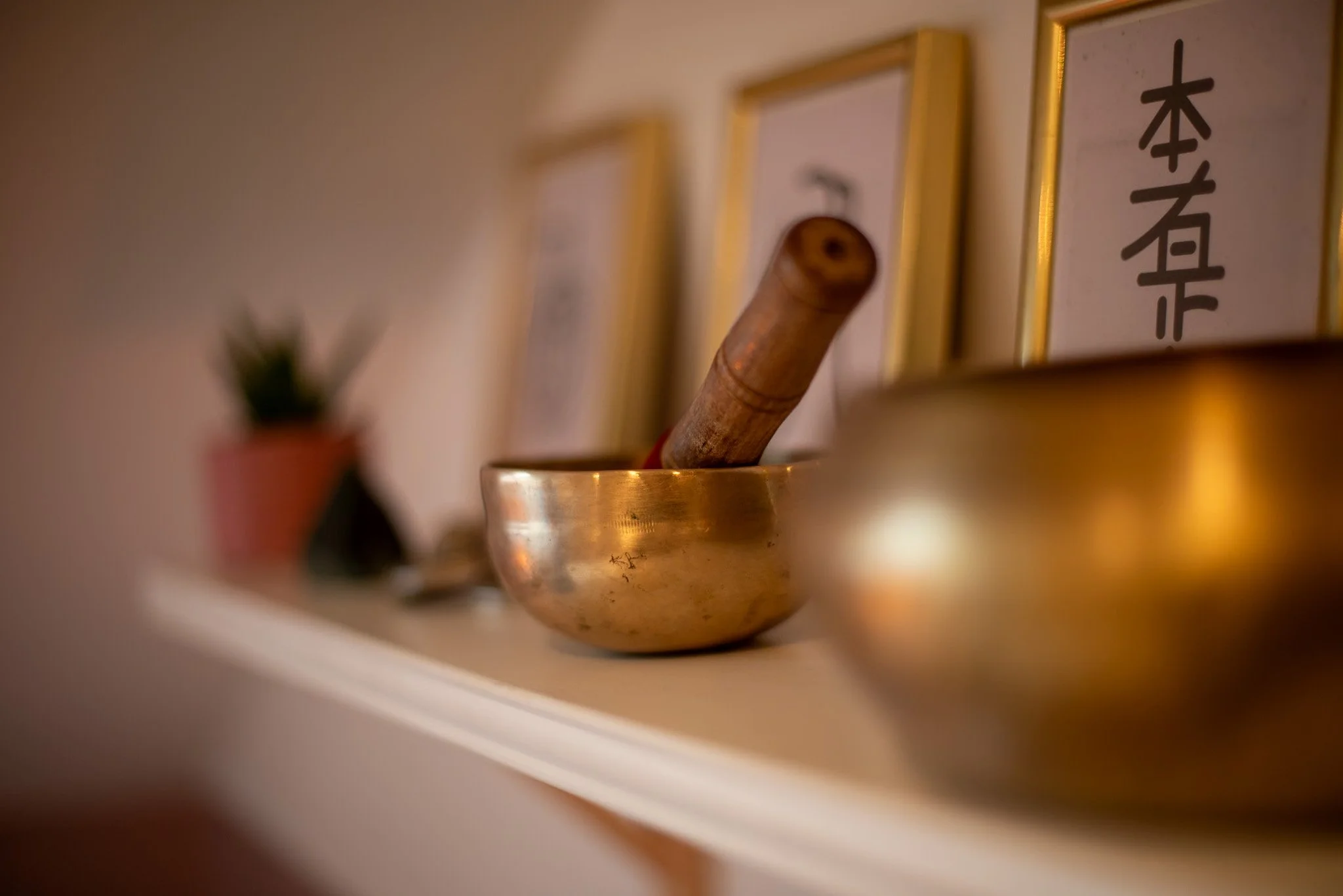 A close-up of a singing bowl with a wooden striker inside, placed on a shelf with framed Reiki symbols in the background and a small potted plant to the left.