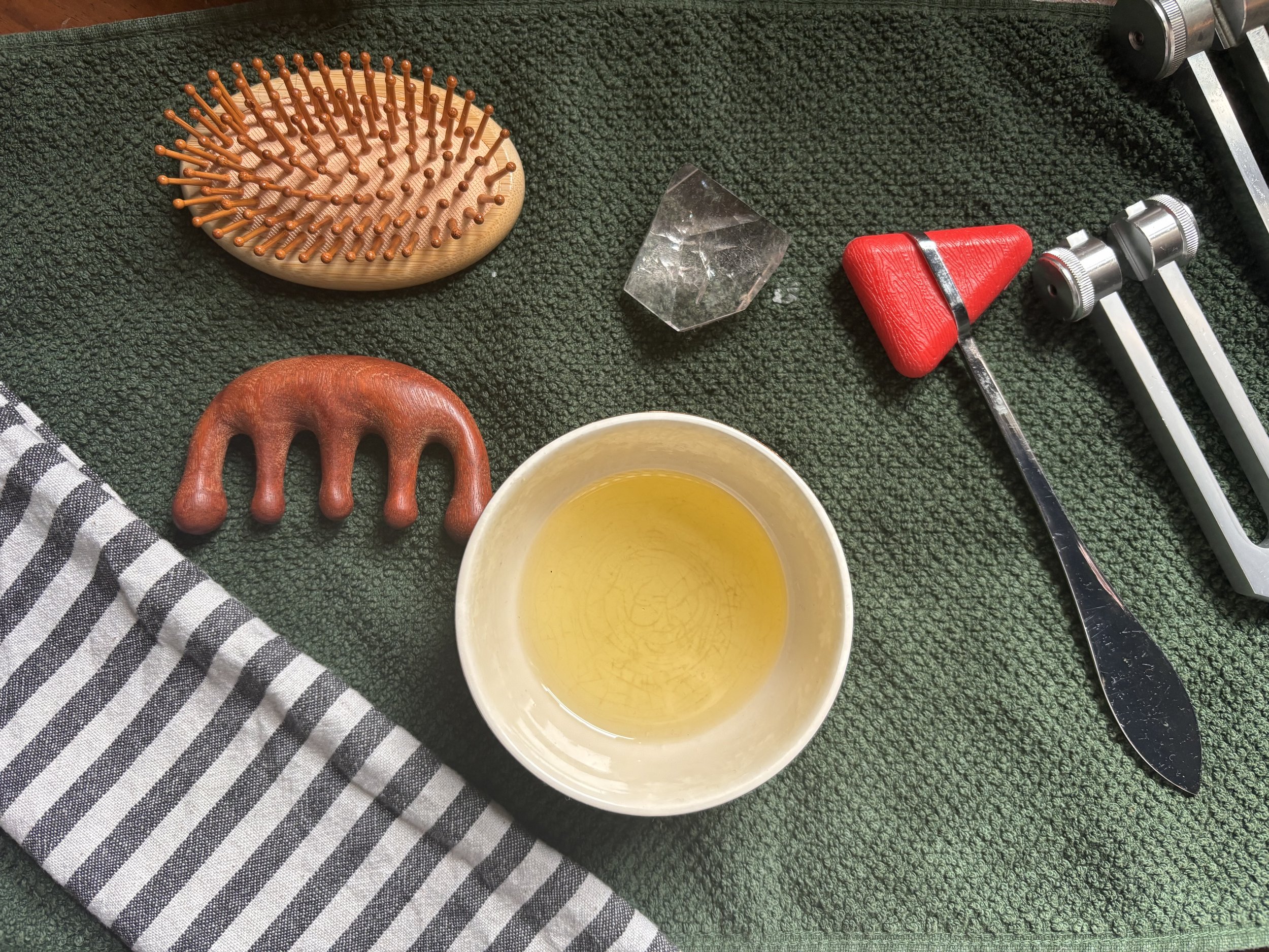 A green textured surface with a black and white eye mask, a wooden hair brush and gua sha tool alongside a clear quartz crustal and weighted tuning fork.