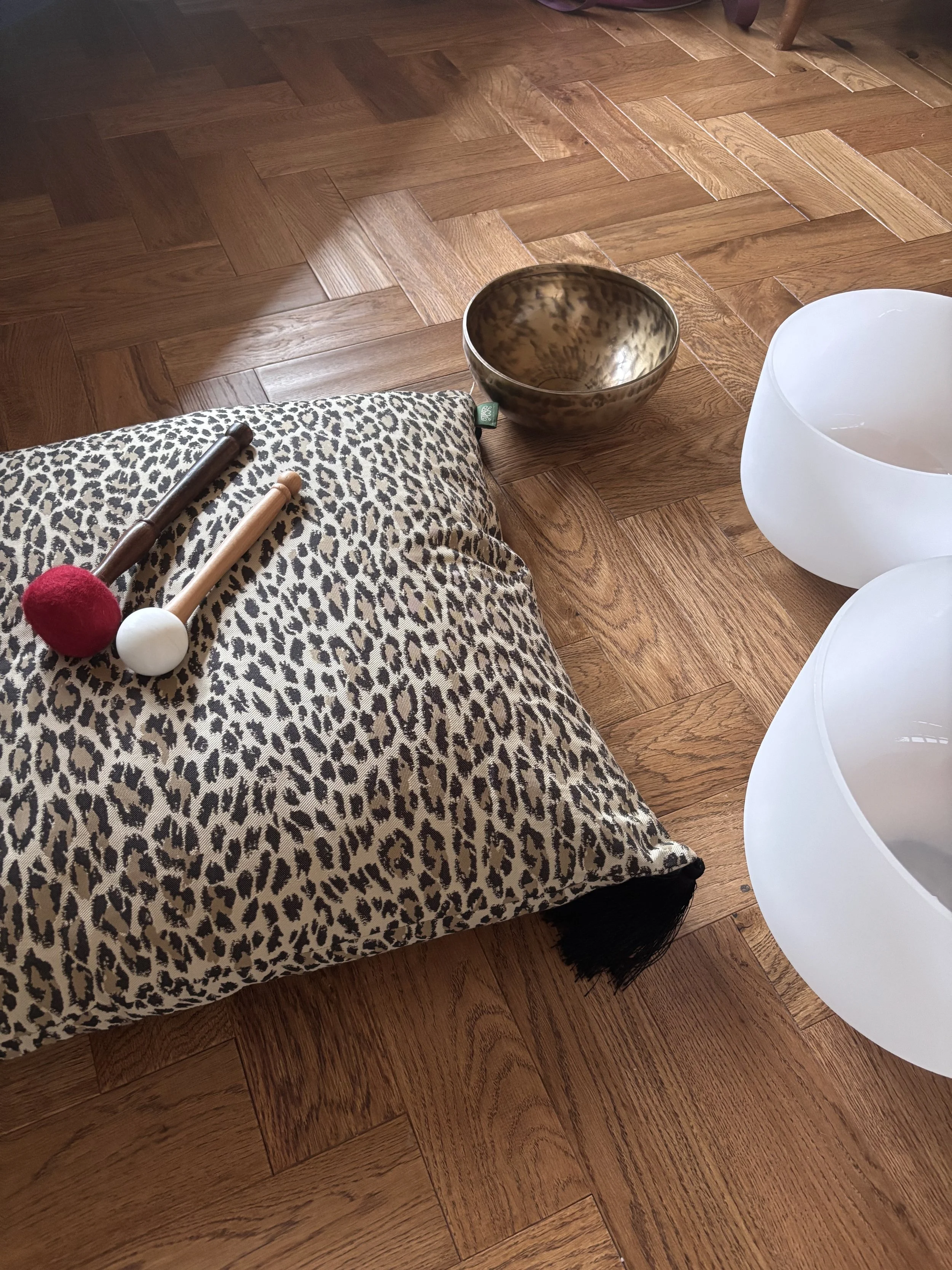 Leopard print cushion on wooden floor with two mallets, a metallic singing bowl, and two white crystal singing bowls.
