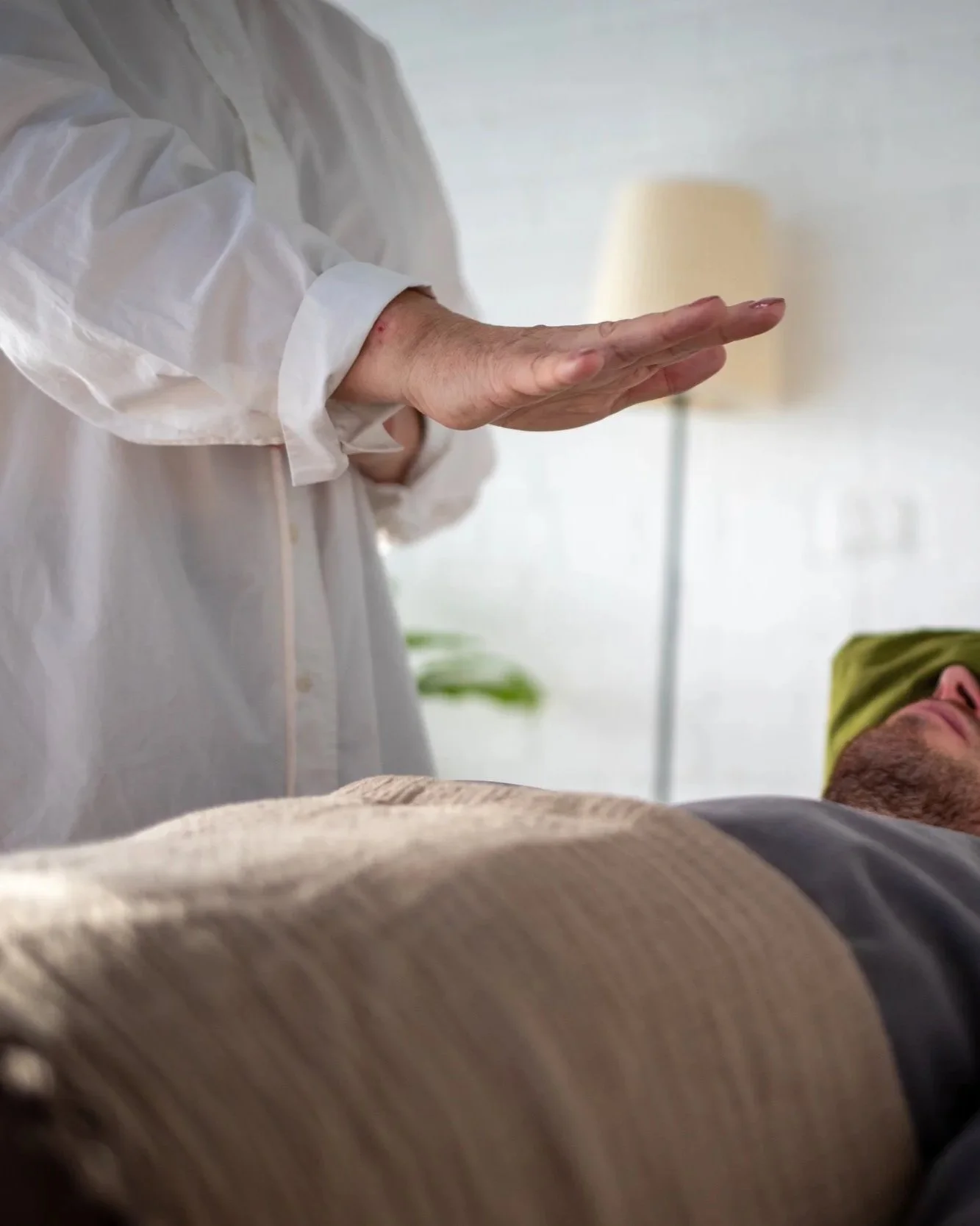 a white woman wearing a white shirt with rolled up sleeves is channeling Reiki energy to a white man lying on a massage bed with a beard