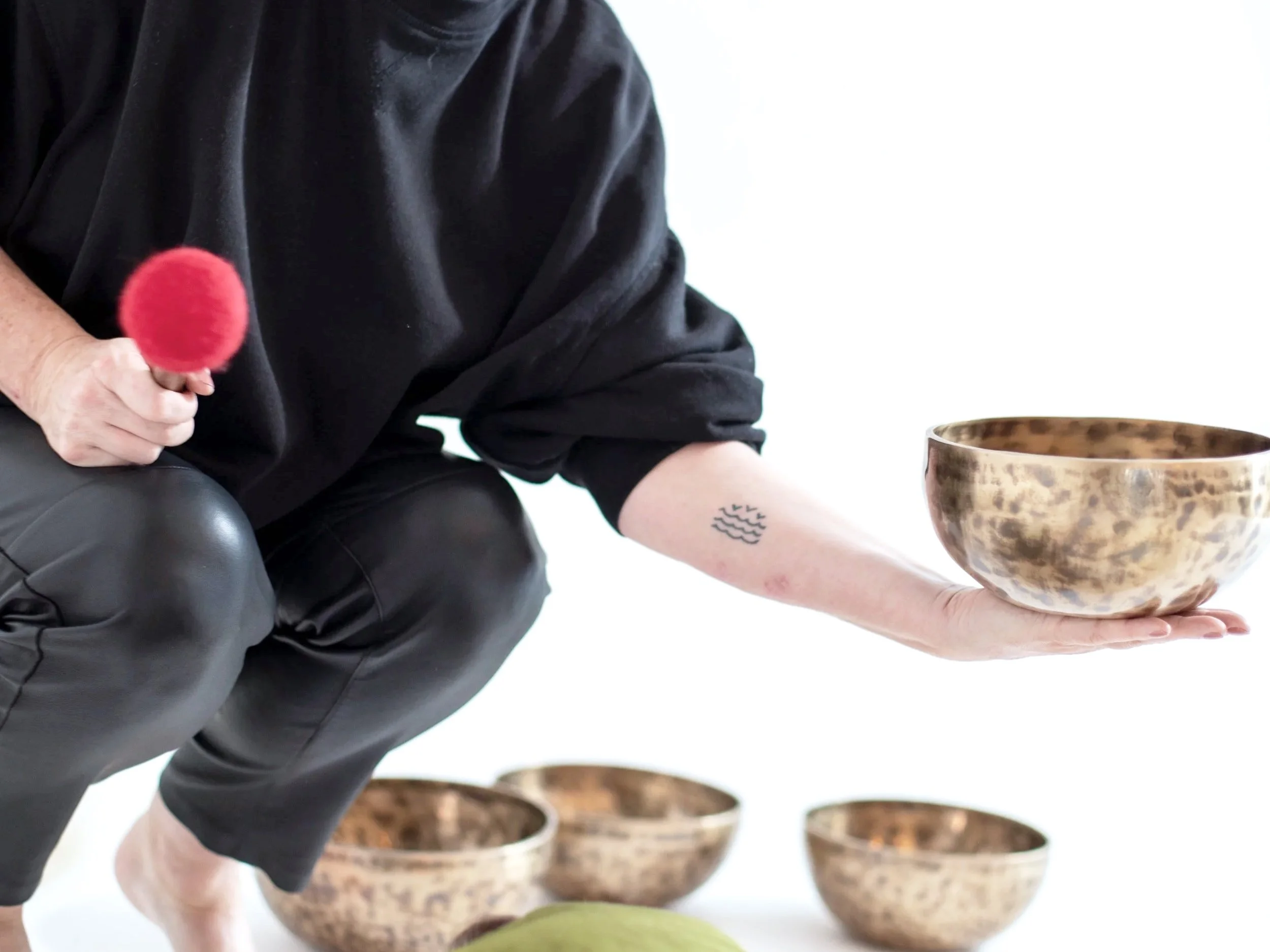 Person crouching, wearing black shirt and pants, holding a red mallet in one hand and a metallic bowl in the other, with other similar bowls in the background.