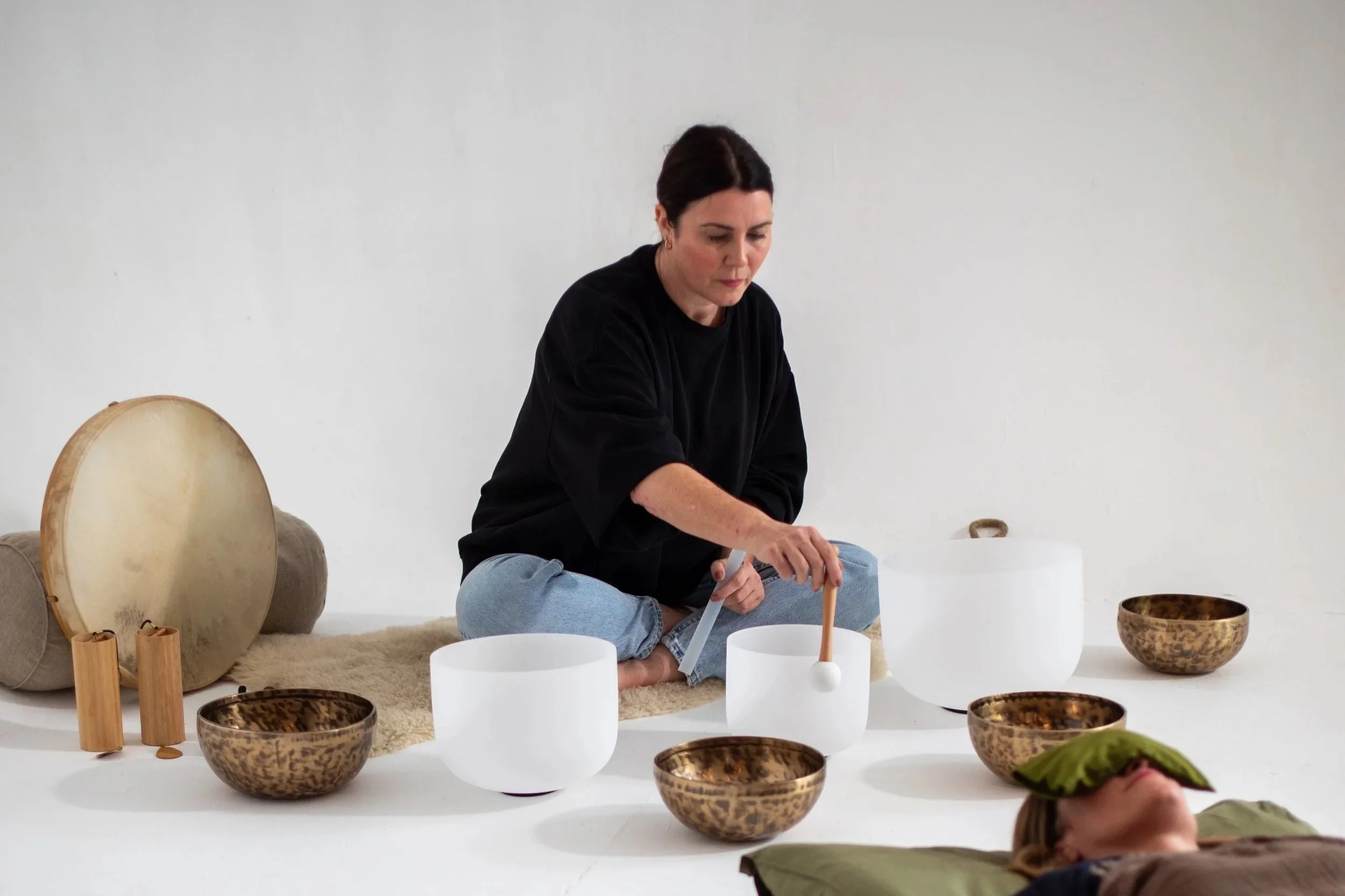 A woman sitting cross-legged on a cream rug surrounded by glass and wooden singing bowls, with a person lying down nearby with a green cloth over their face, in a minimalistic white space.