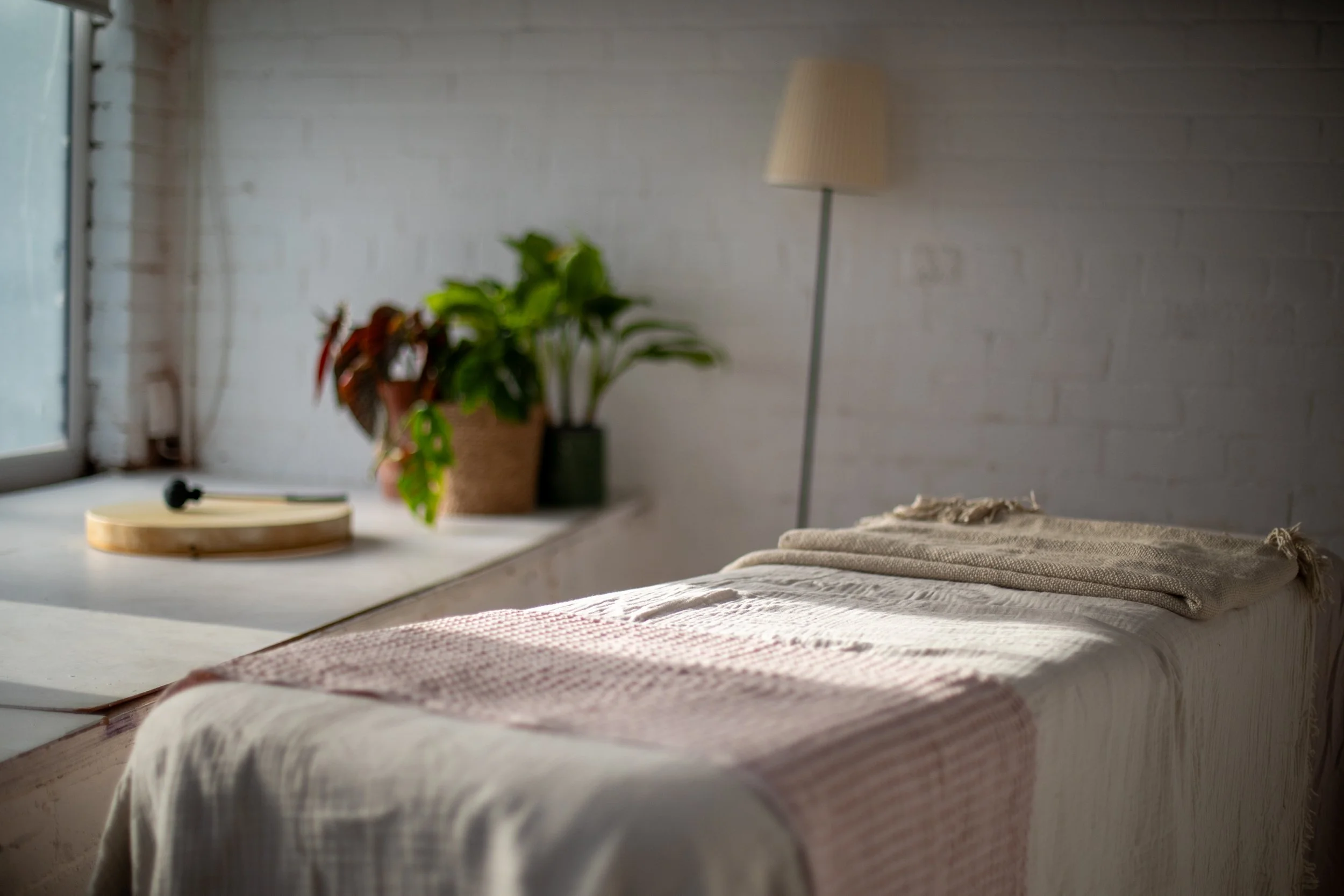 A cozy, sunlit room with a massage bed, a white blanket, a folded towel, a standing lamp, potted plants on the windowsill, a drum and drum beater