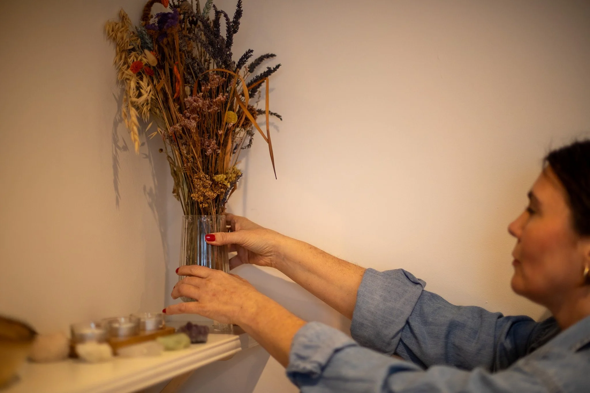 A woman arranging dried flowers in a tall glass vase on a wall shelf.