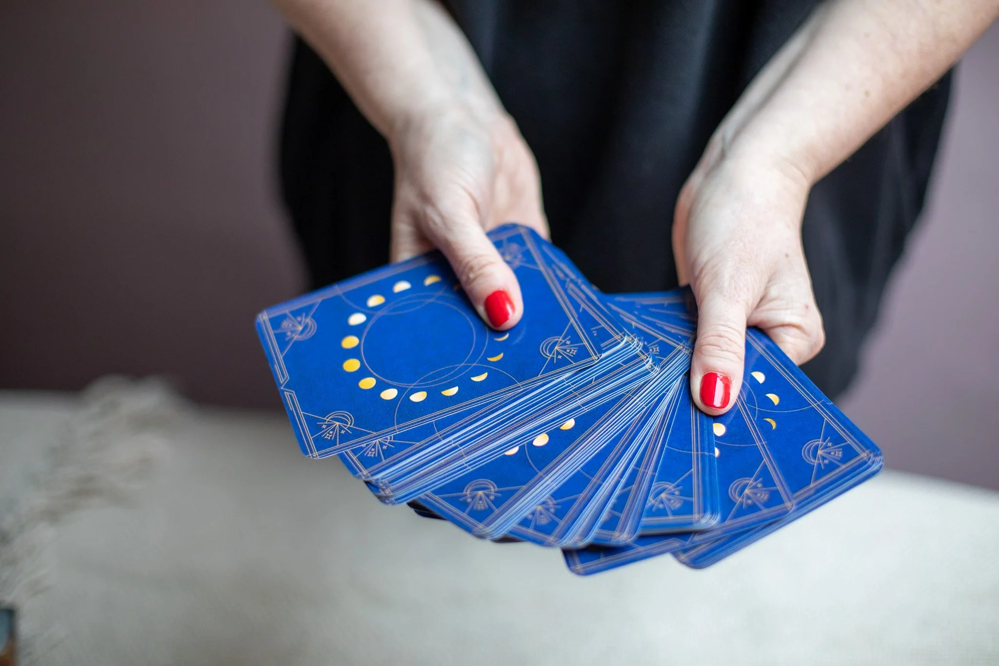 A person with red nail polish holding a fan of blue tarot cards with white and yellow designs.