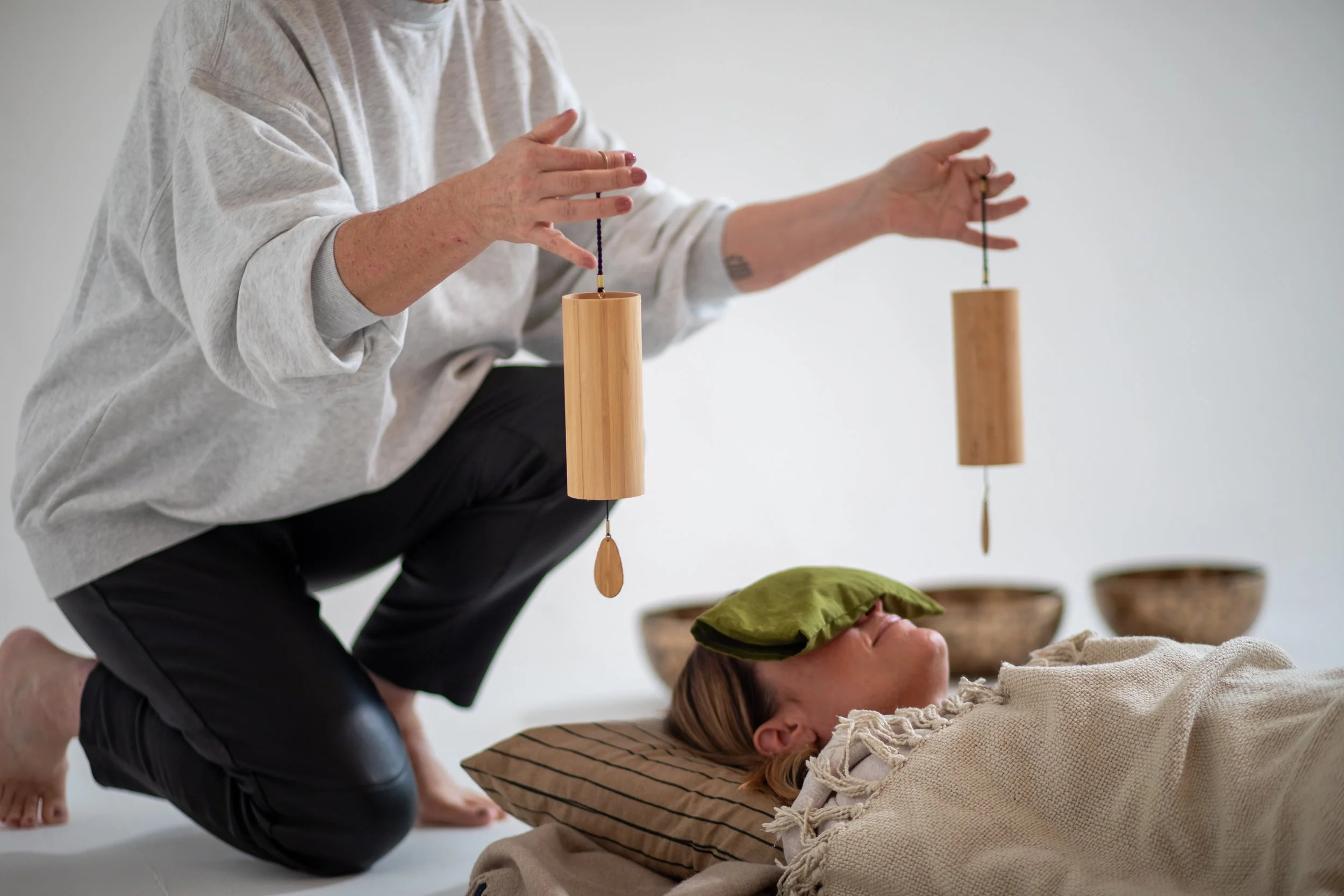 A person is kneeling and holding two wooden sleep meditation chimes over a woman lying down with a green pillow on her face, in a minimalistic room with four metal singing bowl in the background.