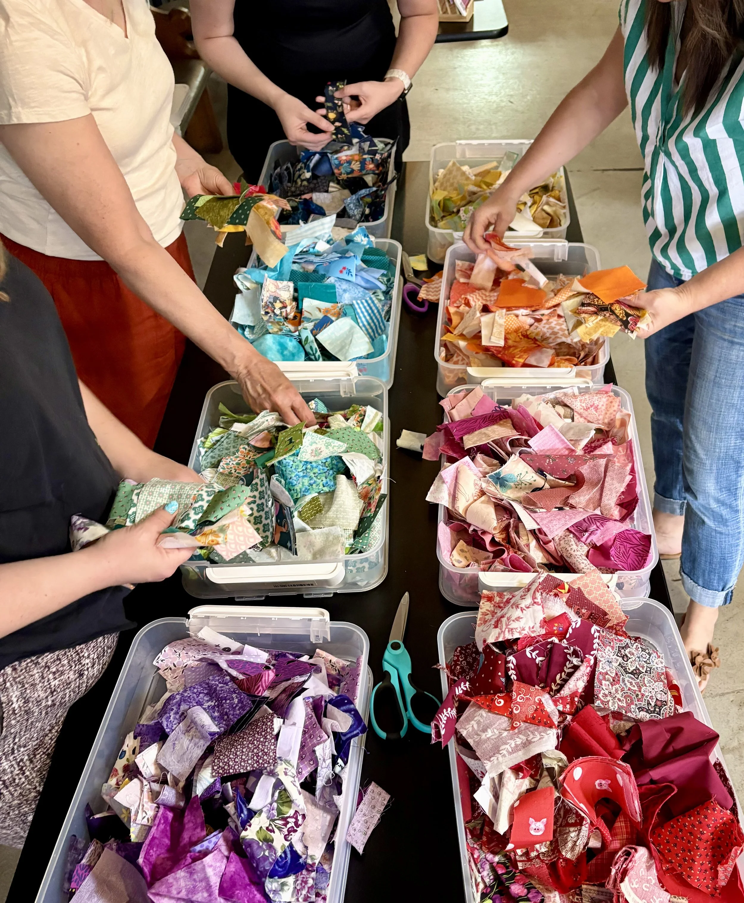 People sorting through colorful fabric scraps in plastic containers, with scissors and scissors on the table.