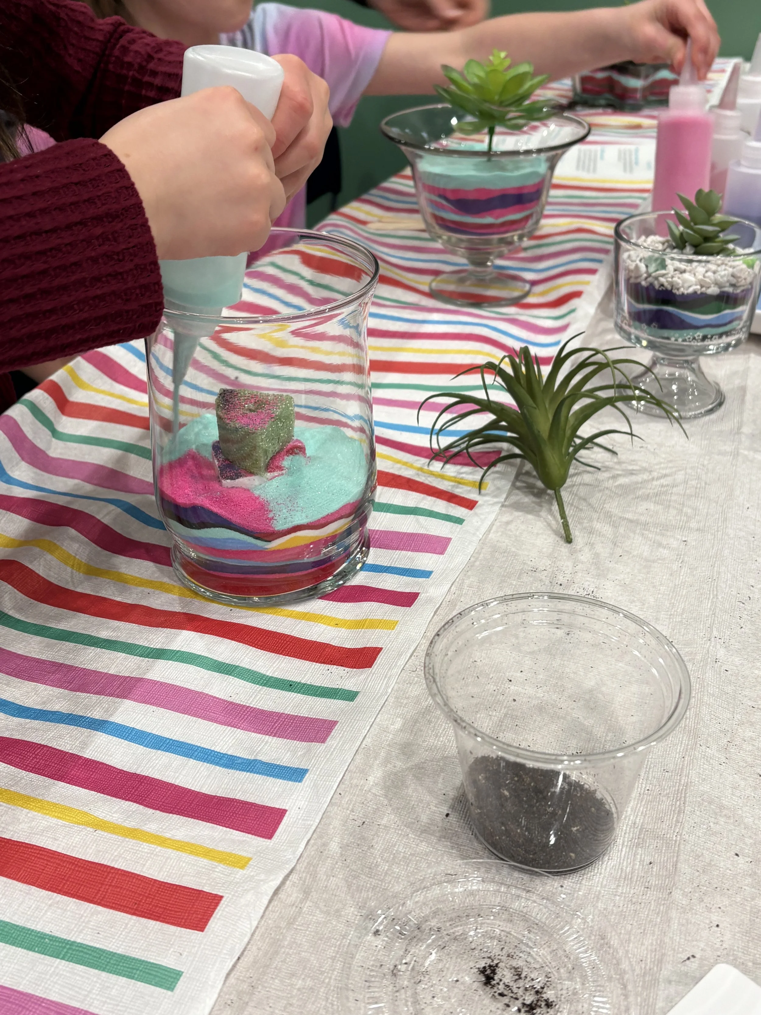 People are pouring colored powder into glass containers on a table decorated with a striped tablecloth. The table also has small potted succulents and other supplies for crafting.
