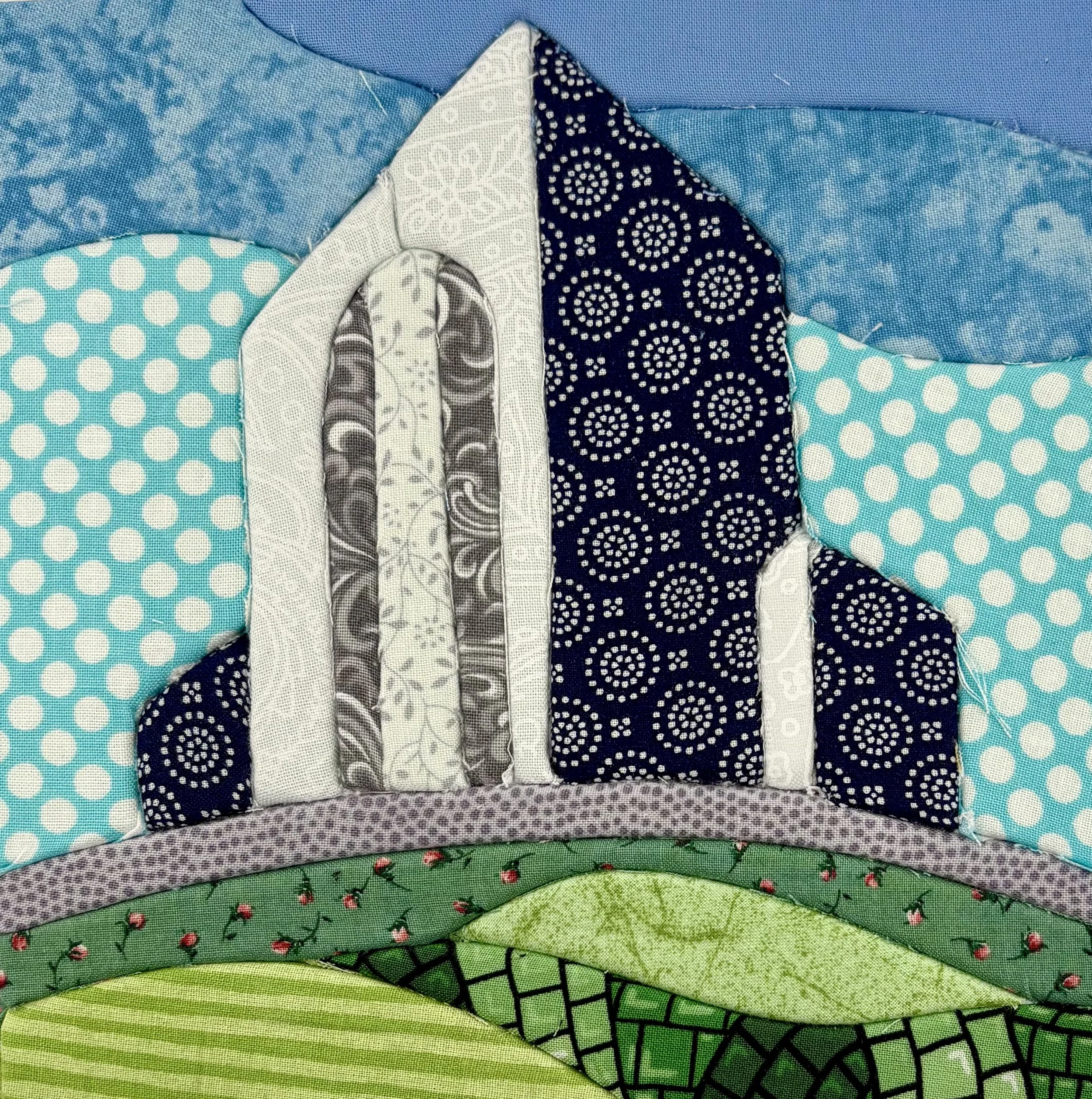 Stacked patchwork quilts in various colors and patterns including blue, white, black, green, and floral designs.