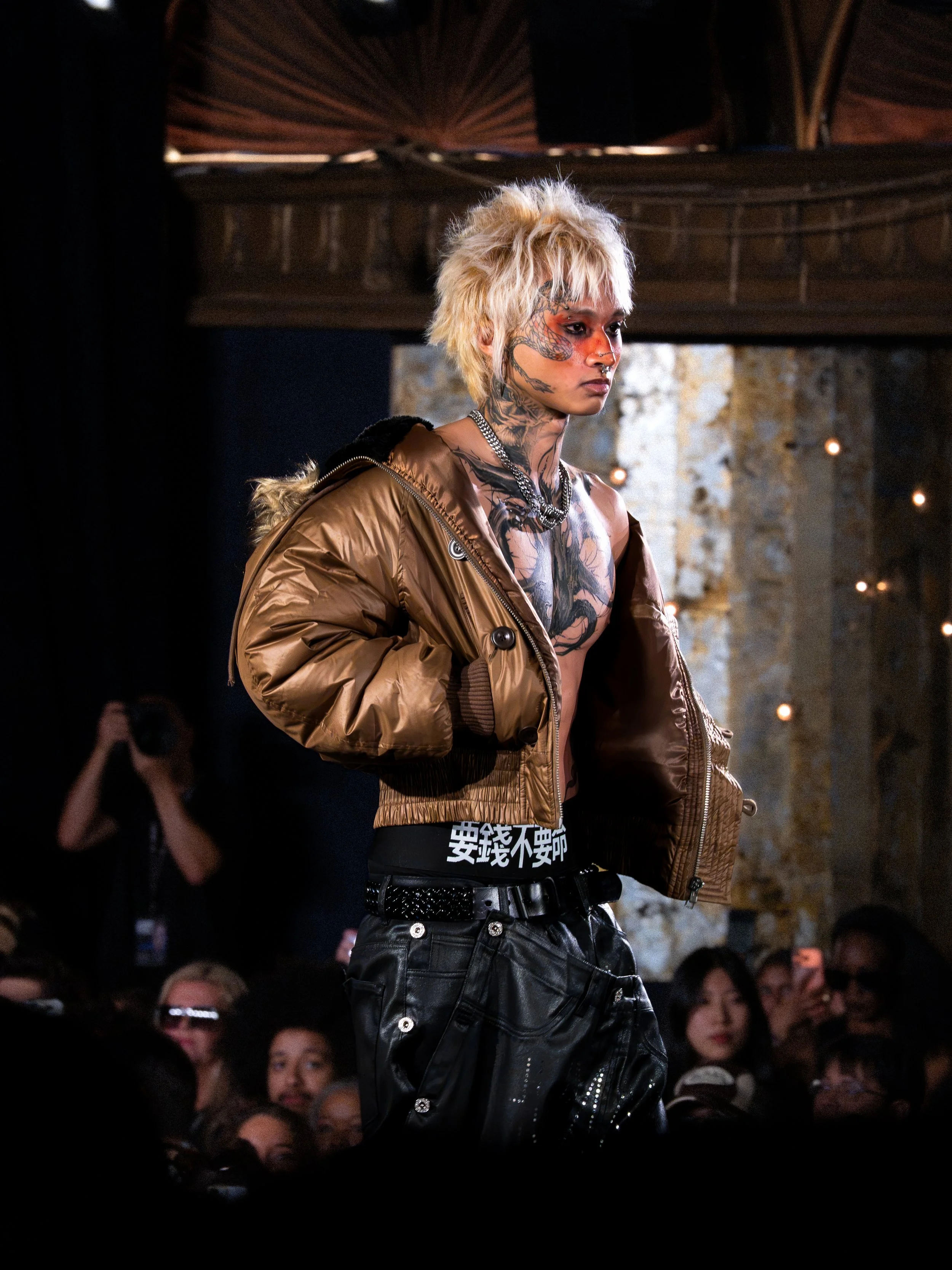 A person with short, bleached blonde hair and extensive tattoos on their face, neck, and chest, standing on a runway during a fashion show. They are wearing a tan bomber jacket and black pants with silver hardware, with a serious expression and audie