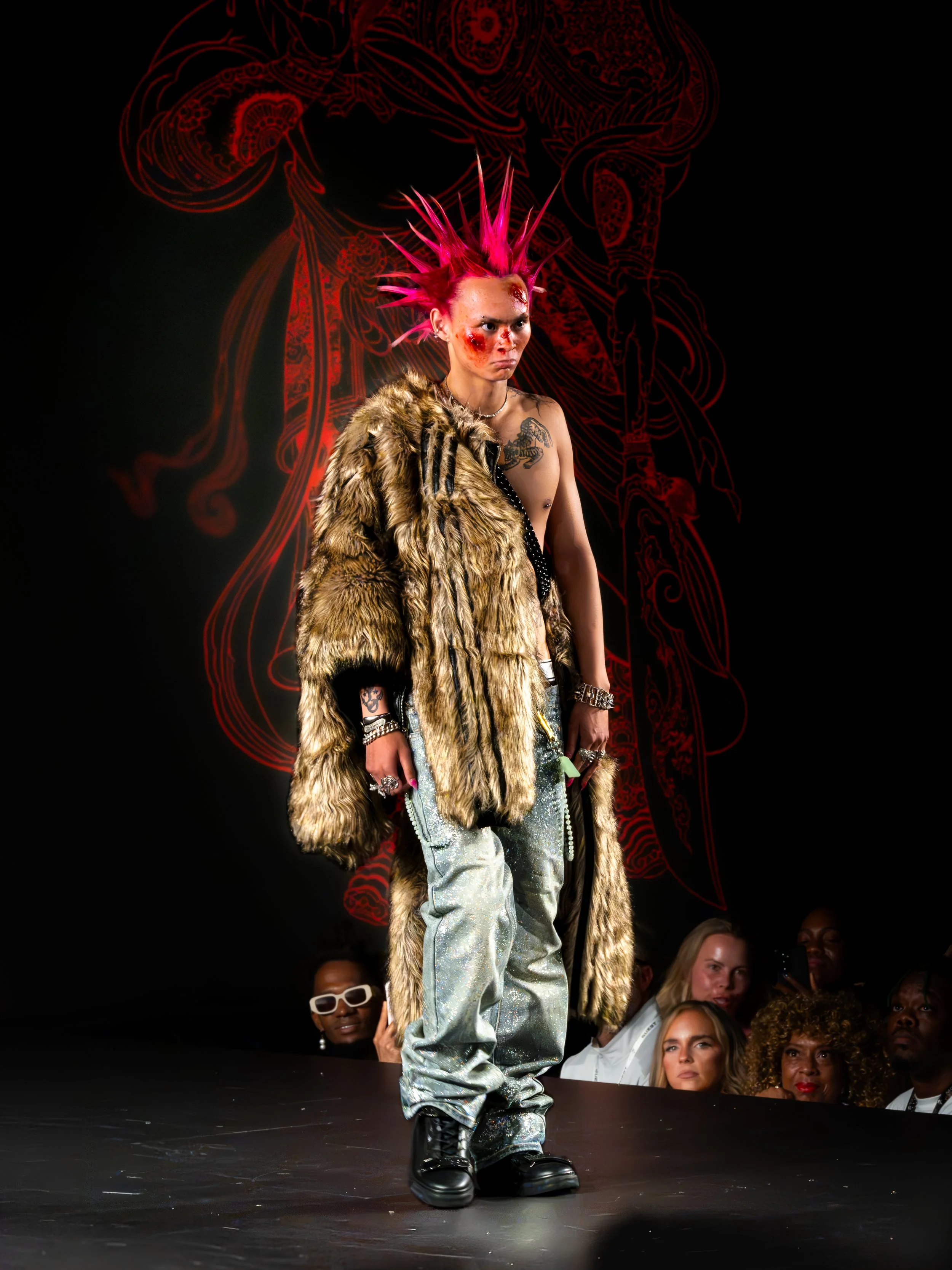 A person with pink spiked hair, makeup with scars, wearing a fur coat, shiny pants, and black boots walking on a runway during a fashion show, with a red outline of a figure in the background and an audience watching.