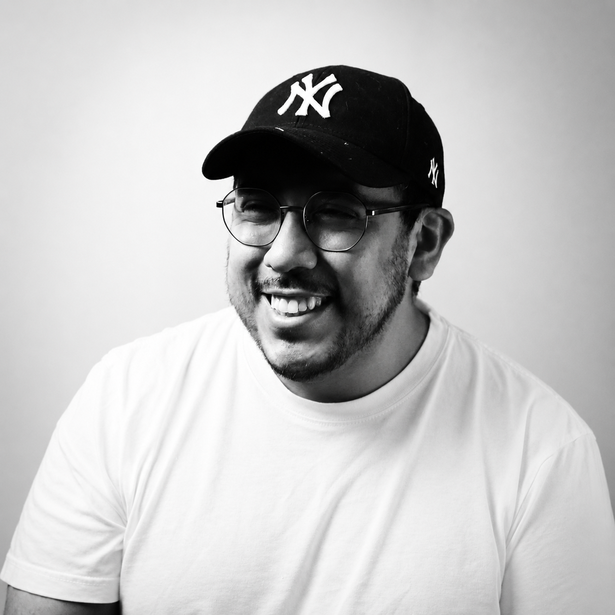 A smiling man wearing glasses and a New York Yankees baseball cap, dressed in a white t-shirt, in front of a plain wall.